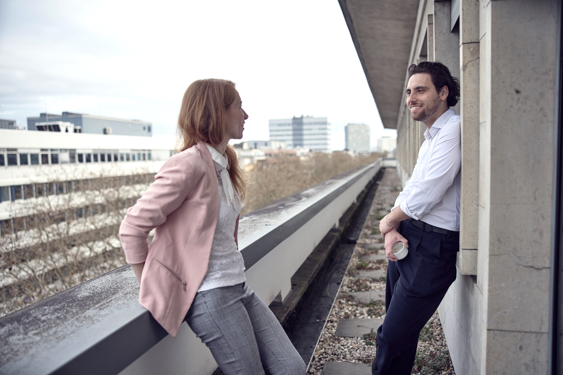 Zwei Personen unterhalten sich entspannt auf einem Balkon, während sie urbanen Hintergrund mit modernen Gebäuden im Hintergrund genießen. Die Frau trägt eine hellrosa Blazerjacke und die Männer einen weißen Hemd, während er eine Tasse in der Hand hält.