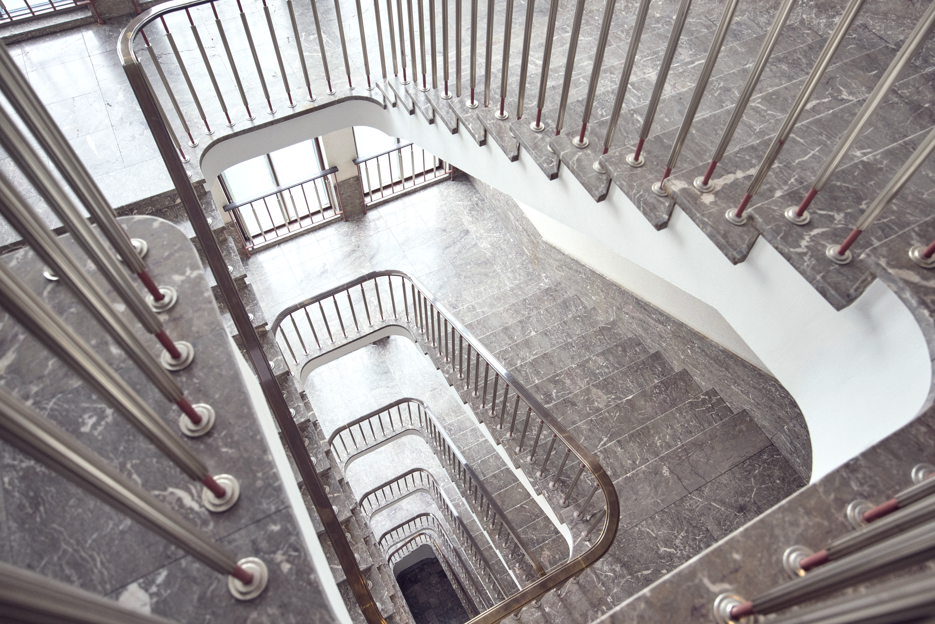 Eine moderne Treppe mit geschwungenen Geländern aus Metall, die in mehreren Ebenen nach unten führt. Die Stufen sind mit einem dunklen, marmorierten Bodenbelag versehen. Durch große Fenster fällt natürliches Licht in den Treppenraum.