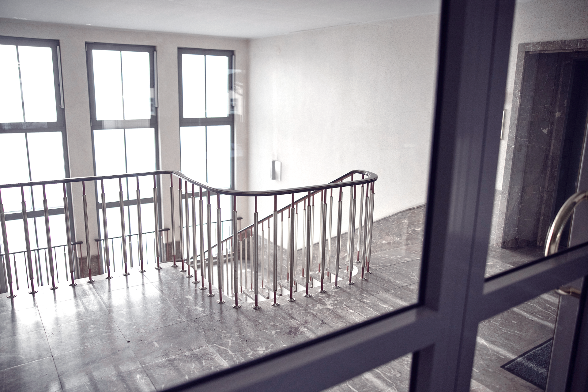 Eine schlichte Treppe mit einem gebogenen Geländer in einem hell erleuchteten Treppenhaus, umgeben von großen Fenstern. Der Boden ist mit grauem Marmor ausgelegt.