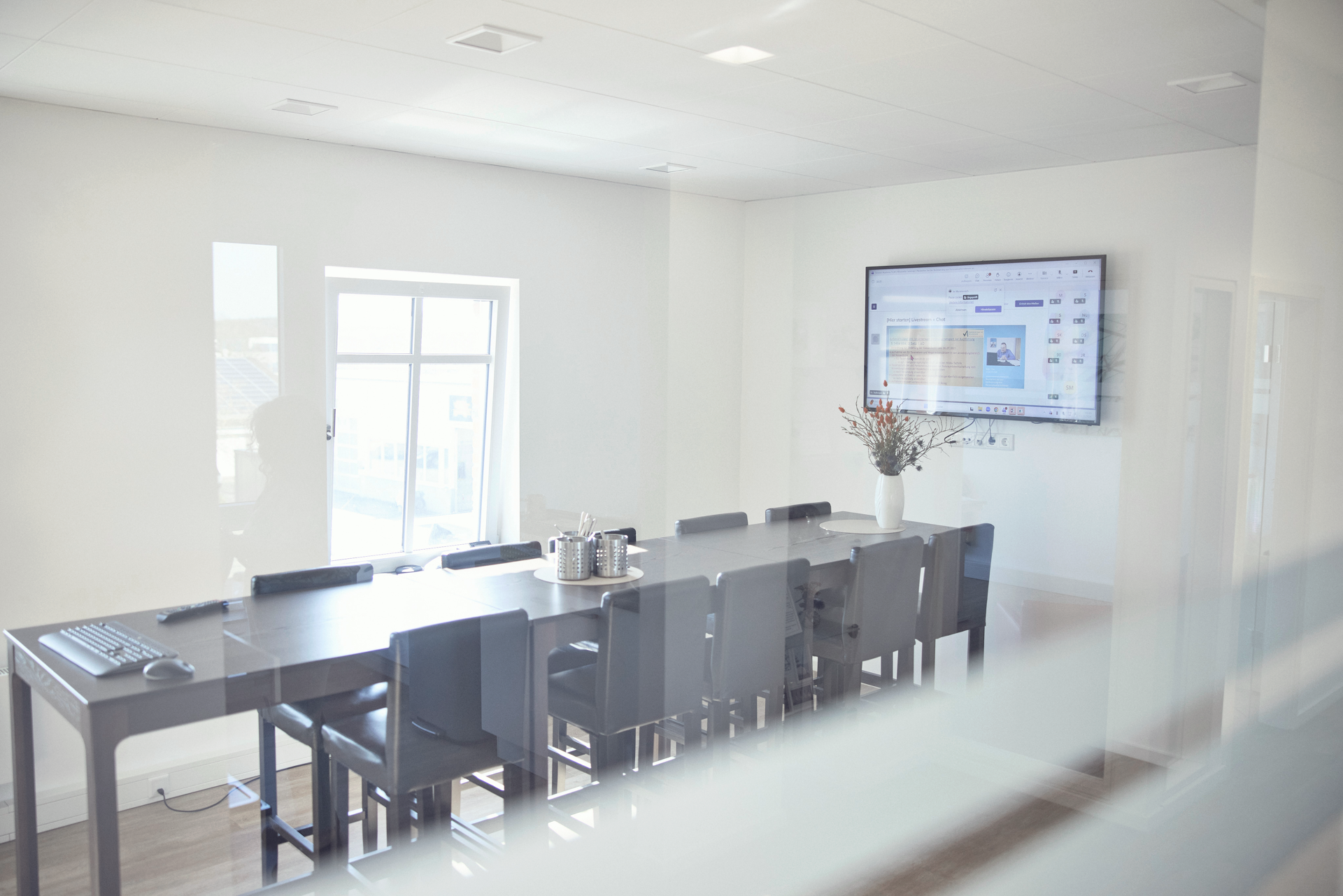 Ein heller, moderner Konferenzraum mit einem langen Tisch und Stühlen, ausgestattet mit einem Monitor an der Wand, auf dem eine Präsentation angezeigt wird. Ein Tisch mit Kaffeekannen und einer Blumenvase ist ebenfalls sichtbar.