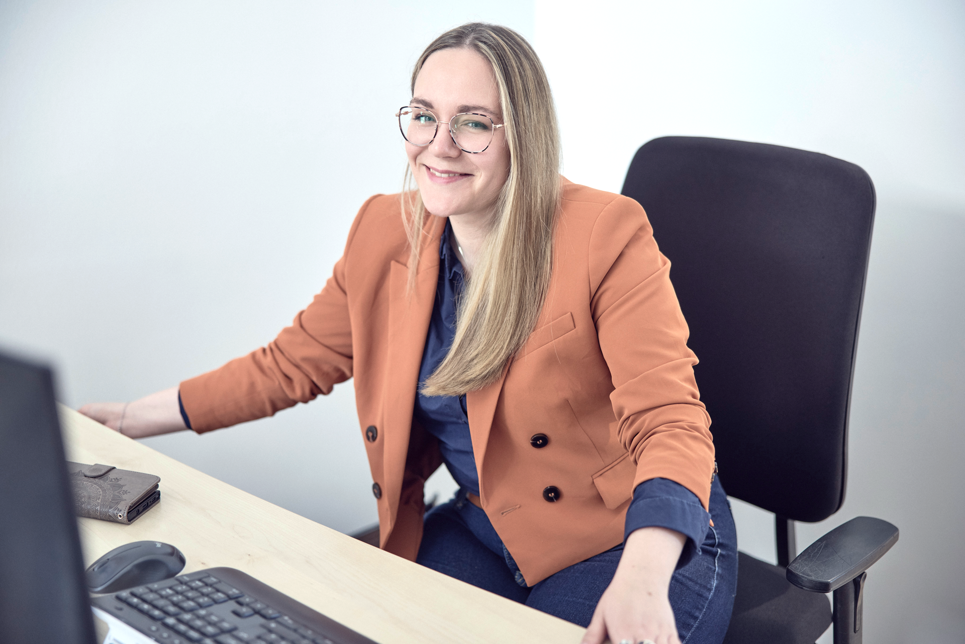 Eine Person mit langen blonden Haaren sitzt an einem Schreibtisch und lächelt in die Kamera. Sie trägt einen orangefarbenen Blazer über einem blauen Oberteil. Am Schreibtisch befinden sich ein Computer und eine Maus.