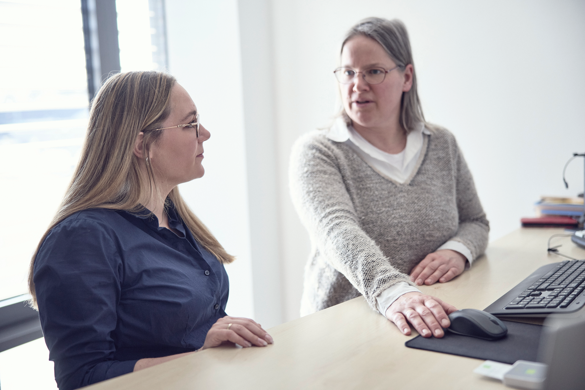 Zwei Frauen sitzen an einem Schreibtisch, wobei eine Frau mit langen, blonden Haaren in einem blauen Hemd die andere Frau mit grauen Haaren in einem hellen Pullover anblickt, während sie auf eine Maus und einen Computerbildschirm zeigen.