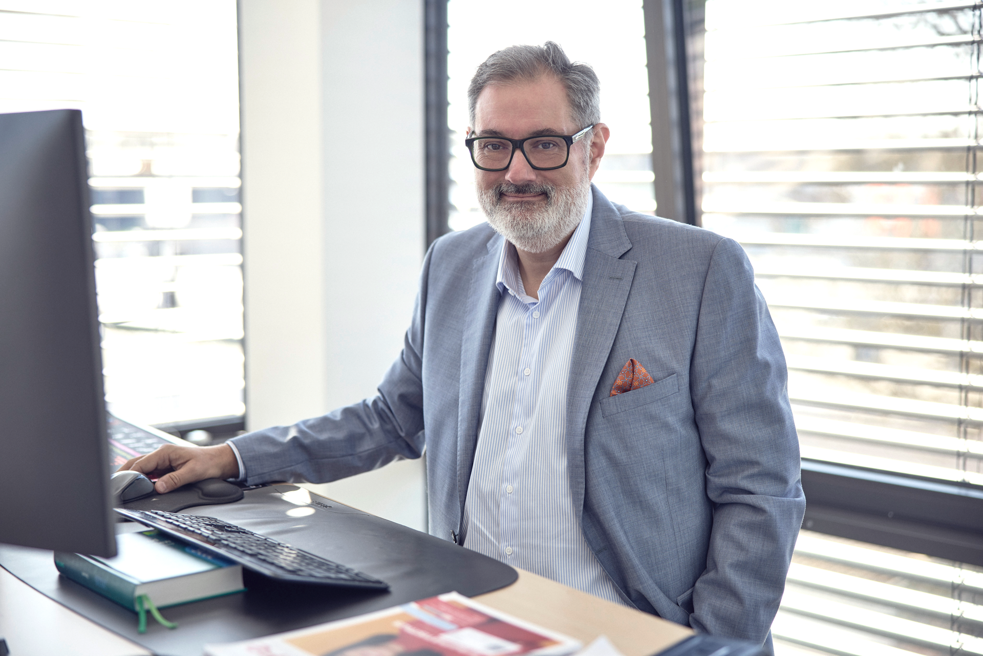 Ein Mann mit Brille und grauem Anzug sitzt an einem Schreibtisch mit Computer in einem modern gestalteten Büro, während er in die Kamera lächelt. Im Hintergrund sind Fenster mit horizontalen Jalousien sichtbar.