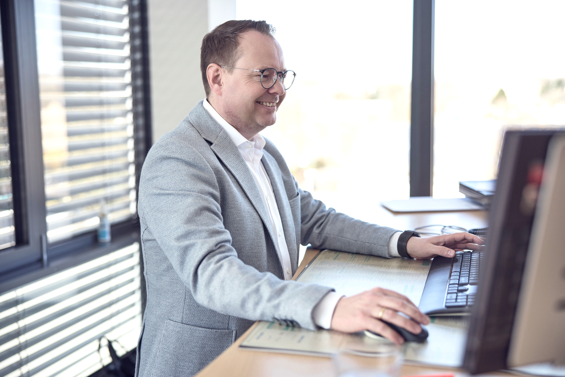 Ein Mann mit Brille sitzt an einem Schreibtisch, arbeitet an einem Computer und lächelt, während er auf die Tastatur schaut. Im Hintergrund sind Fenster mit Blick auf eine Stadtlandschaft.