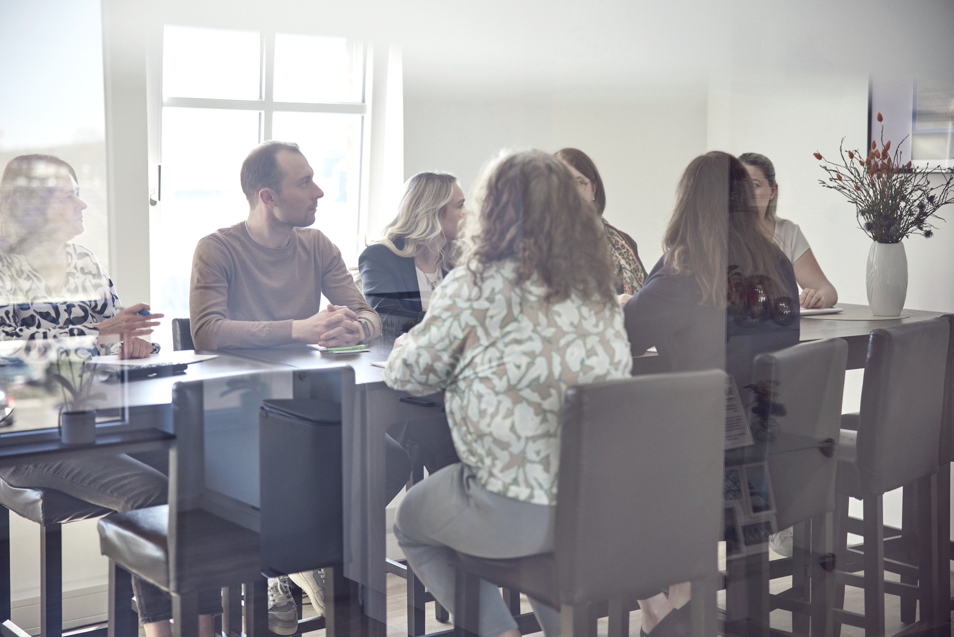 Eine Gruppe von fünf Personen sitzt an einem Tisch in einem hellen Raum, umgeben von Fenstern und einer Pflanze in einer Vase. Sie sind in ein Gespräch vertieft, während eine Person am Kopf des Tisches den anderen zuhört.