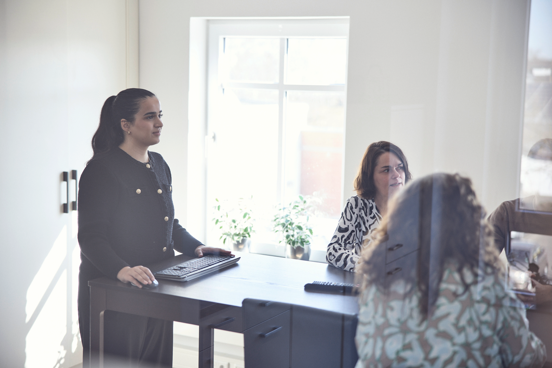 Eine Frau steht an einem Tisch mit einem Laptop, während zwei weitere Frauen am Tisch sitzen und diskutieren. Im Hintergrund sind Pflanzen und ein Fenster zu sehen, das Tageslicht hereinlässt.