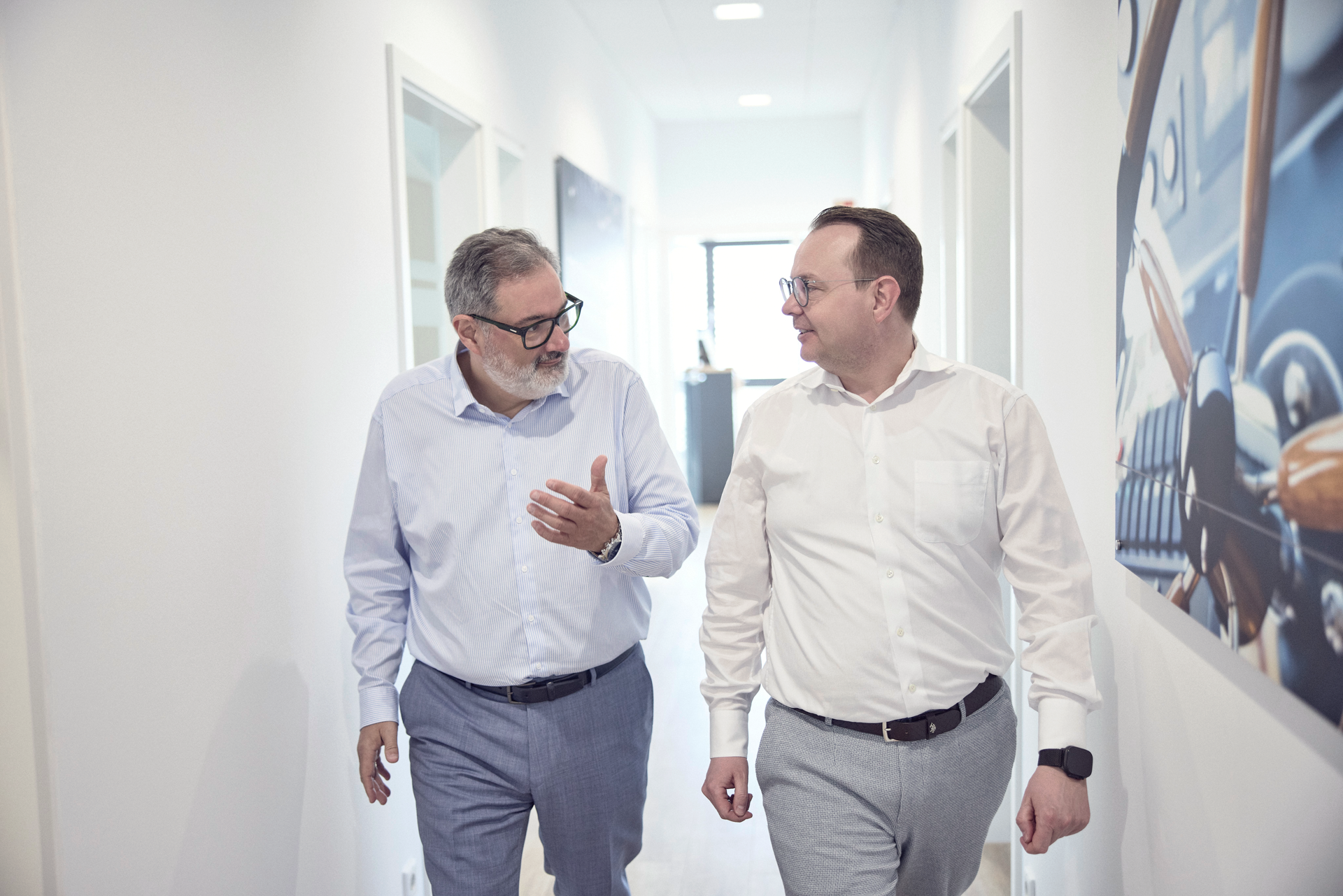 Zwei Männer in formeller Kleidung gehen durch einen hellen Büroflur und sprechen miteinander. Im Hintergrund sind Wandbilder und Bürotüren sichtbar.