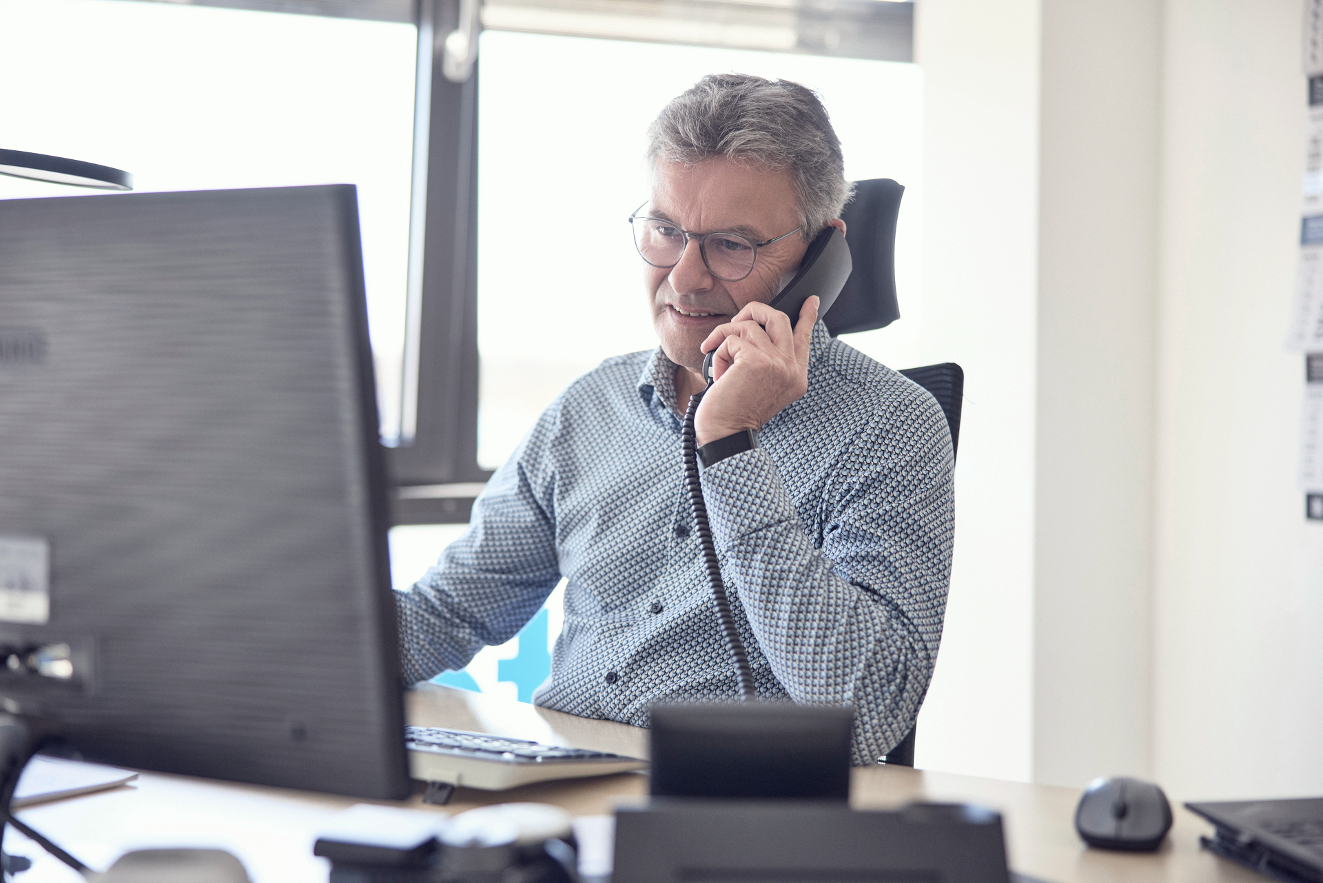 Ein Mann mit Brille und kurzem grauem Haar führt ein Telefongespräch, während er an einem Schreibtisch sitzt und auf einen Computerbildschirm schaut.