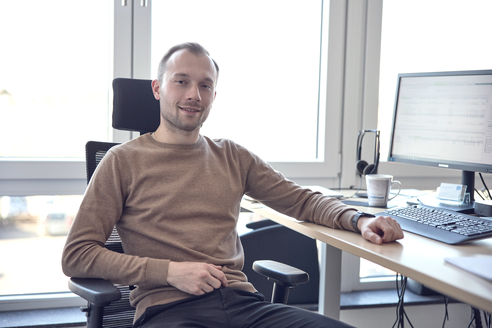 Ein Mann sitzt entspannt in einem Bürostuhl vor einem Schreibtisch mit einem Computerbildschirm, er trägt einen braunen Pullover und lächelt in die Kamera. Im Hintergrund sind Fenster mit Blick auf eine leicht verschwommene Außenansicht zu sehen.