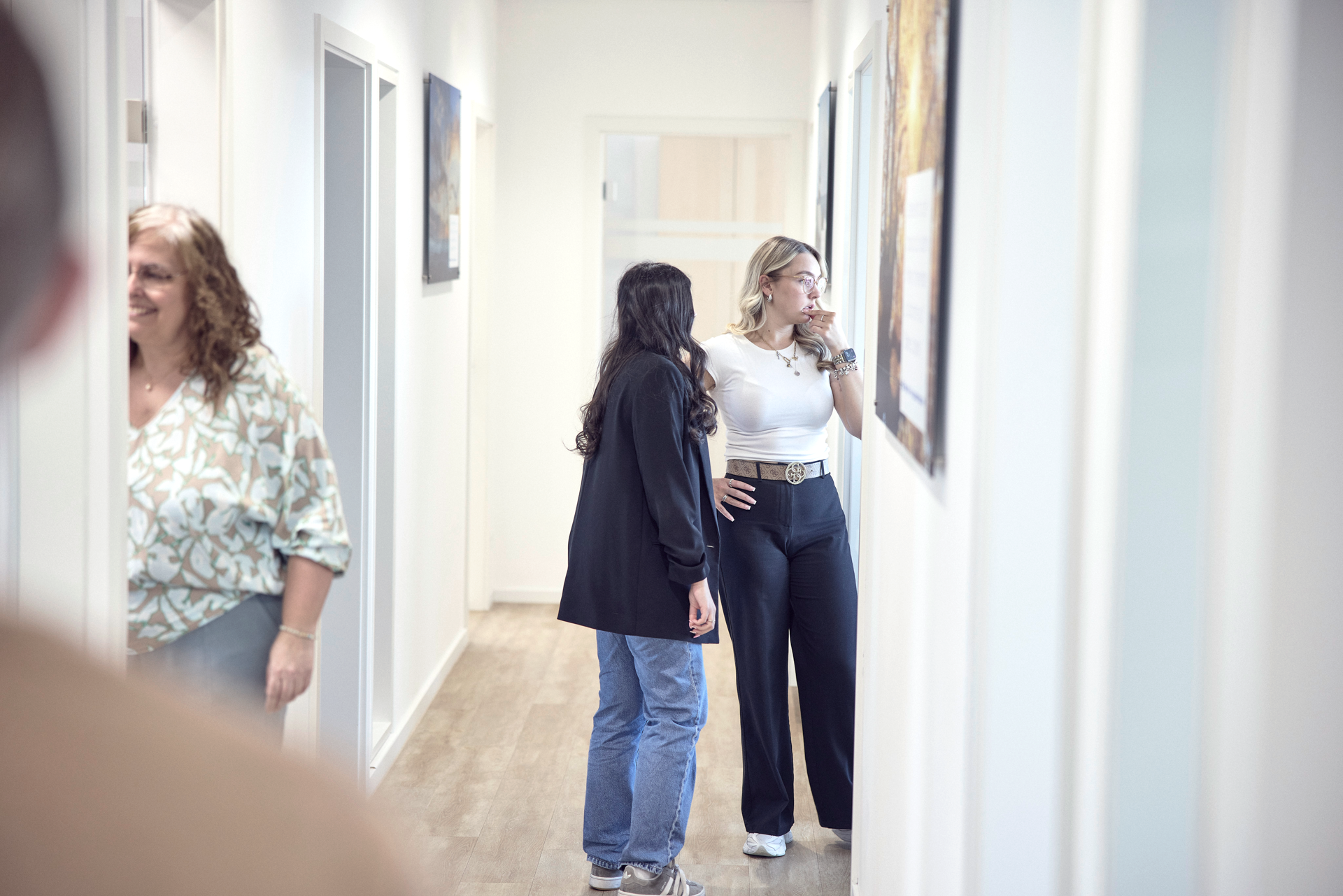 Zwei Frauen stehen in einem hellen Flur eines Gebäudes. Eine Frau trägt eine weiße Bluse und schwarze Hose, während die andere in einem lässigen Outfit mit Jeans und schwarzem Blazer bekleidet ist. Im Hintergrund ist eine dritte Person teilweise sichtbar. An den Wänden hängen Bilder.