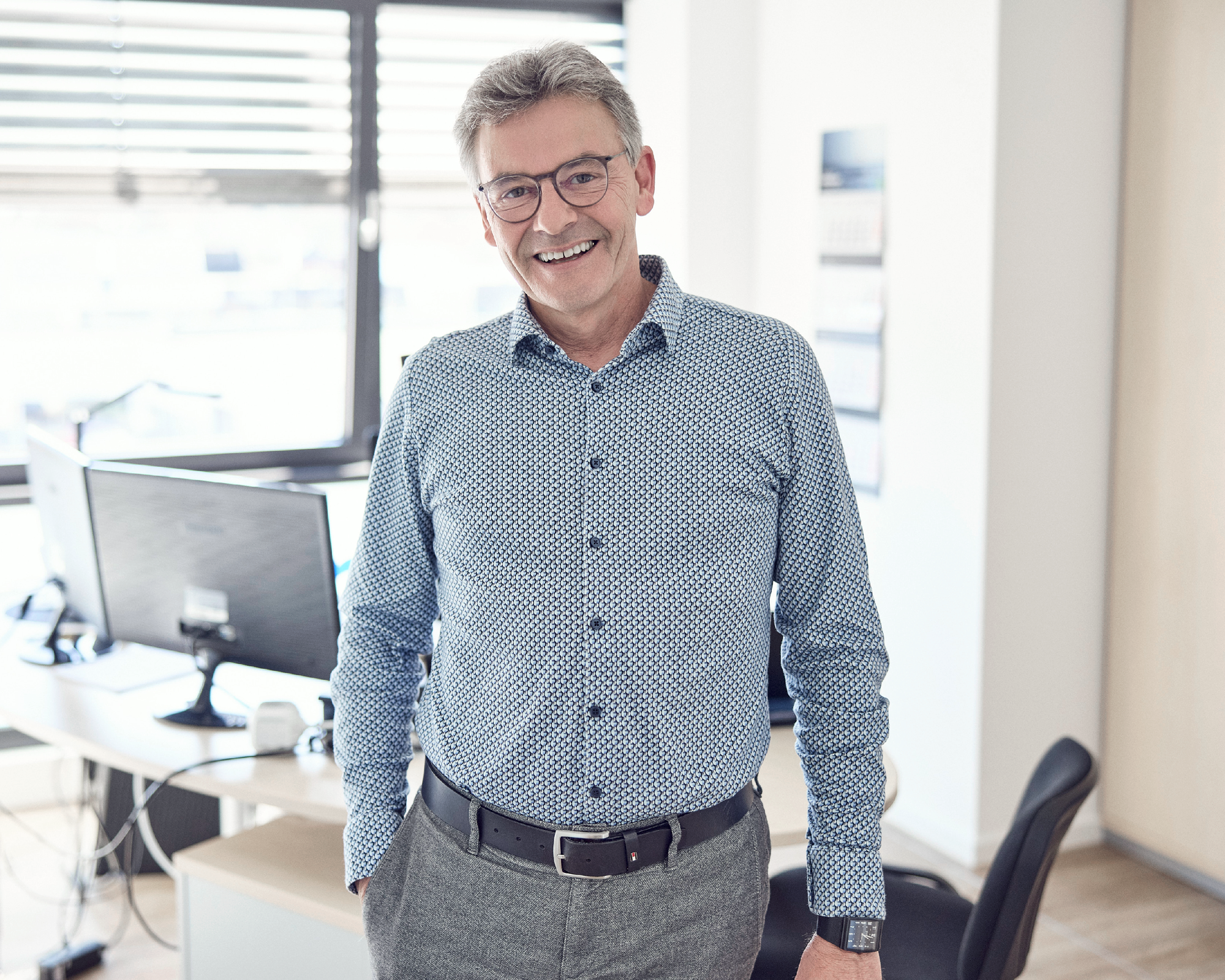 Ein lächelnder Mann mit Brille steht in einem hellen Büro, hinter ihm sind Computer und Büromöbel zu sehen. Er trägt ein blaues, gemustertes Hemd und graue Hosen.