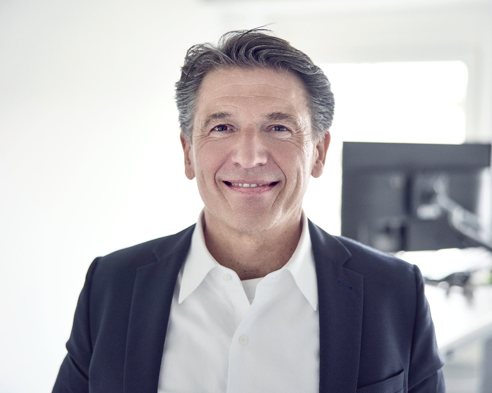 Ein lächelnder Mann mit grauen Haaren trägt einen dunkelblauen Blazer und ein weißes Hemd. Er steht in einem Büro, im Hintergrund ist ein Bildschirm sichtbar.