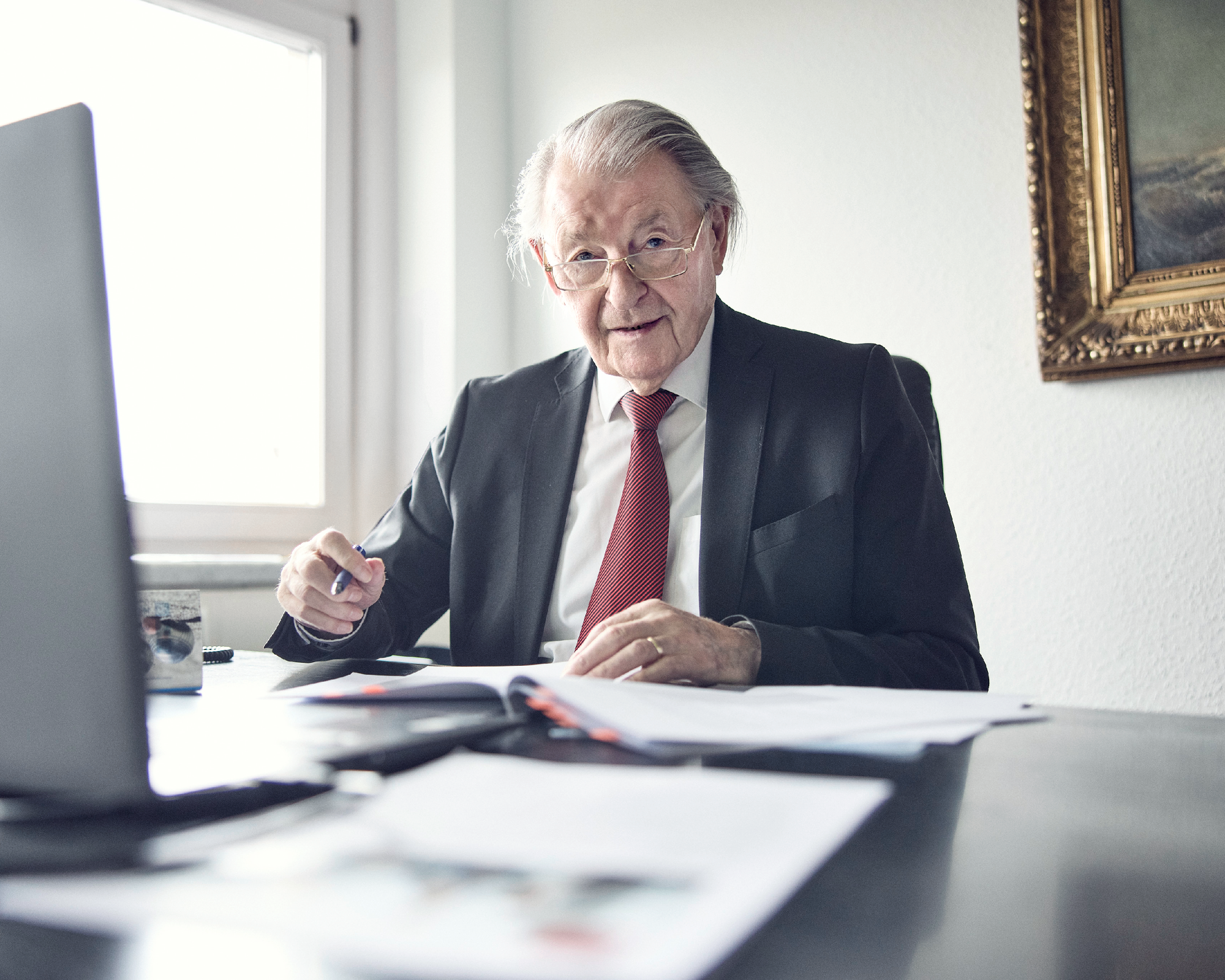 Ein älterer Mann mit Brille und grauen Haaren sitzt an einem Schreibtisch, trägt einen schwarzen Anzug und eine rote Krawatte. Er schaut direkt in die Kamera und hat einen Stift in der Hand, während Dokumente vor ihm auf dem Tisch liegen. Im Hintergrund ist ein Fenster und ein eingerahmtes Bild zu sehen.