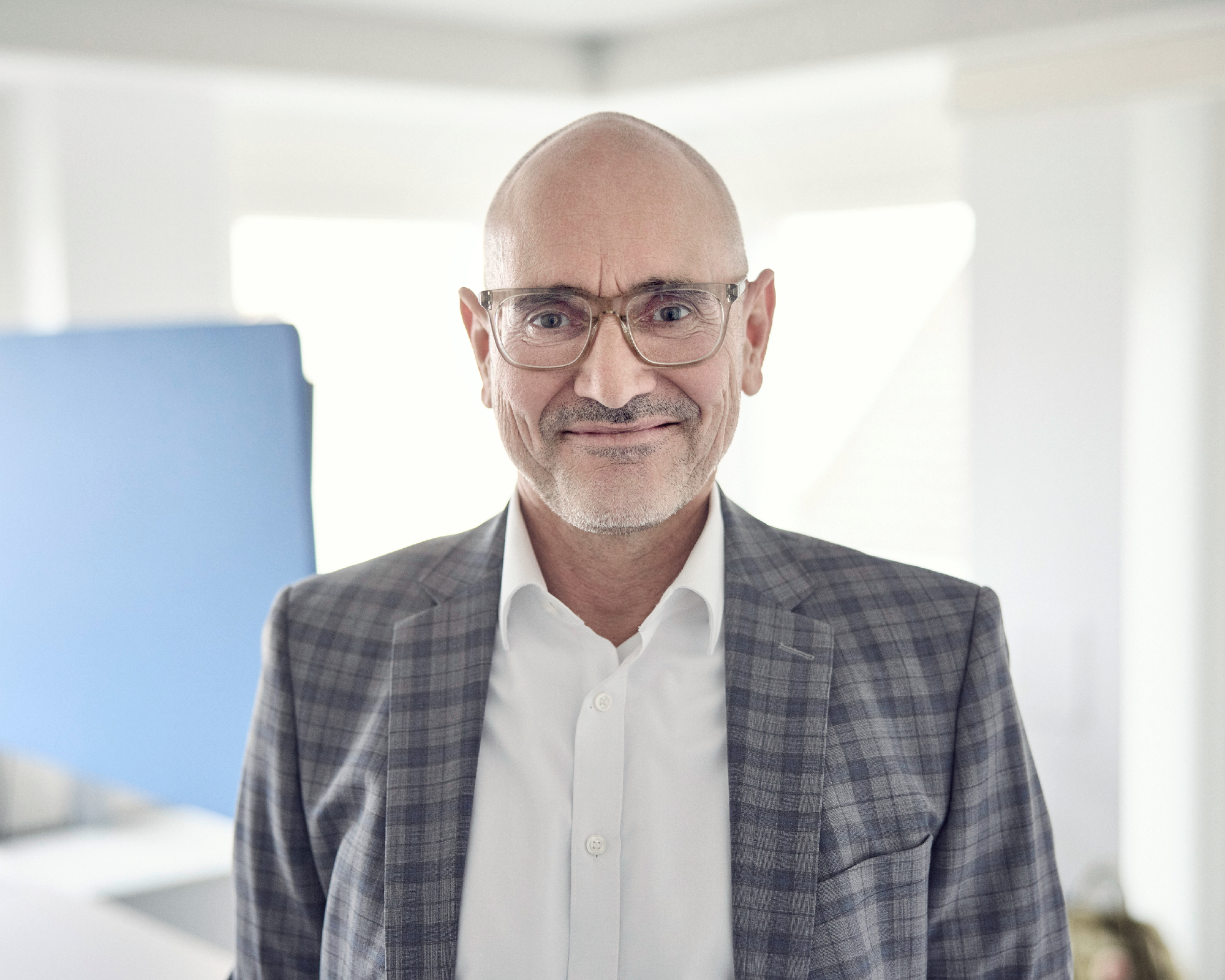 Ein lächelnder Mann mittleren Alters trägt eine Brille und einen karierten Anzug und steht in einem hellen, modernen Raum mit einem blauen Hintergrund.
