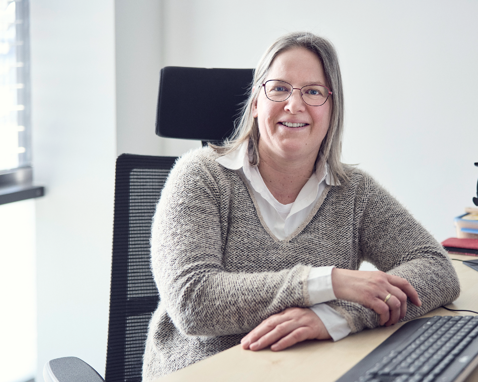 Eine lächelnde Frau mit Brille sitzt an einem Schreibtisch, auf dem ein Computer und einige Bücher liegen. Sie trägt einen beigen Pullover über einem weißen Hemd und hat ihr Haar offen.