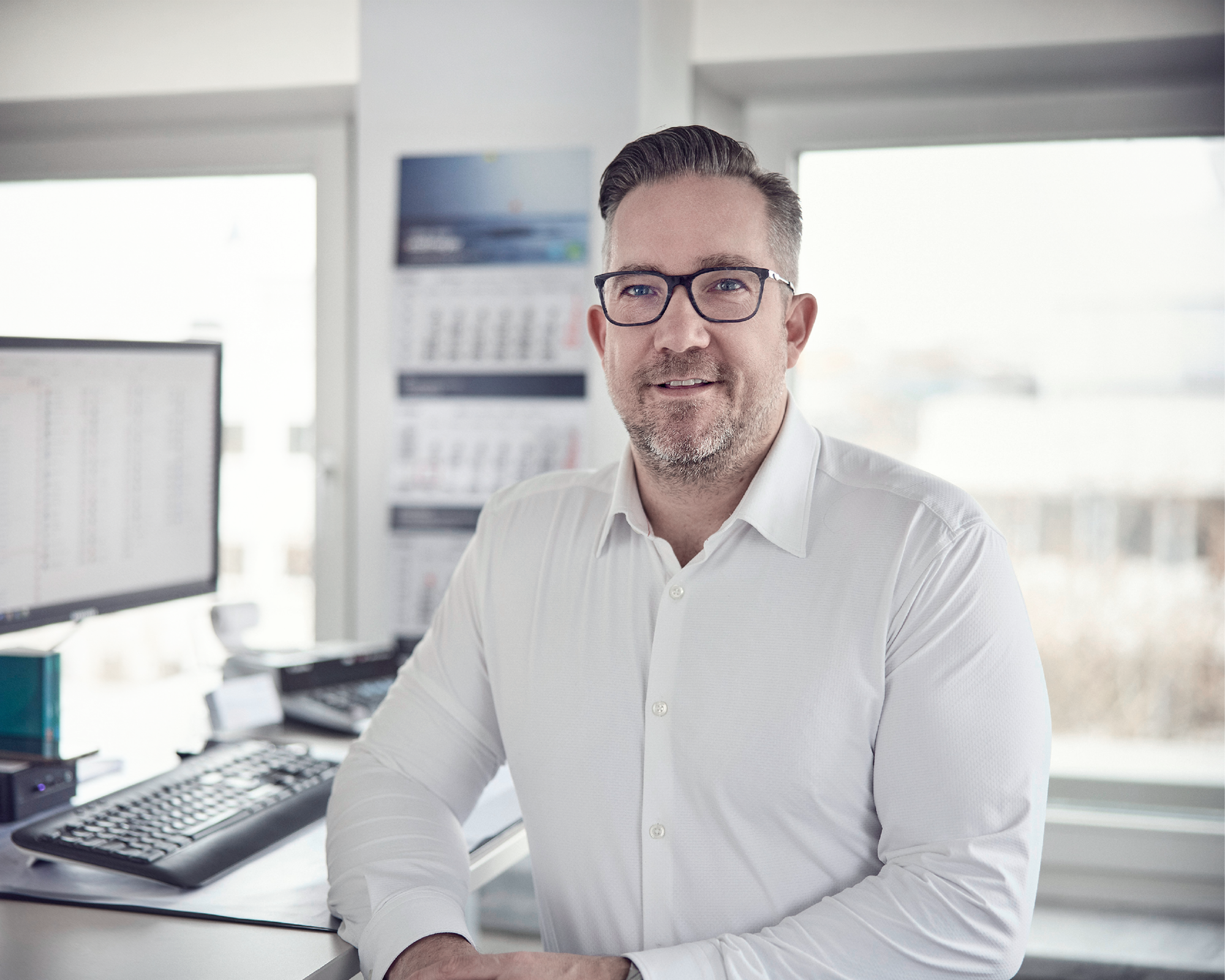 Ein Mann mit Brille und einem weißen Hemd steht in einem Büro, während er an einem Schreibtisch mit einem Computer und einer Tastatur lehnt, im Hintergrund sind Fenster mit Tageslicht und eine Wand mit einem Kalender zu sehen.