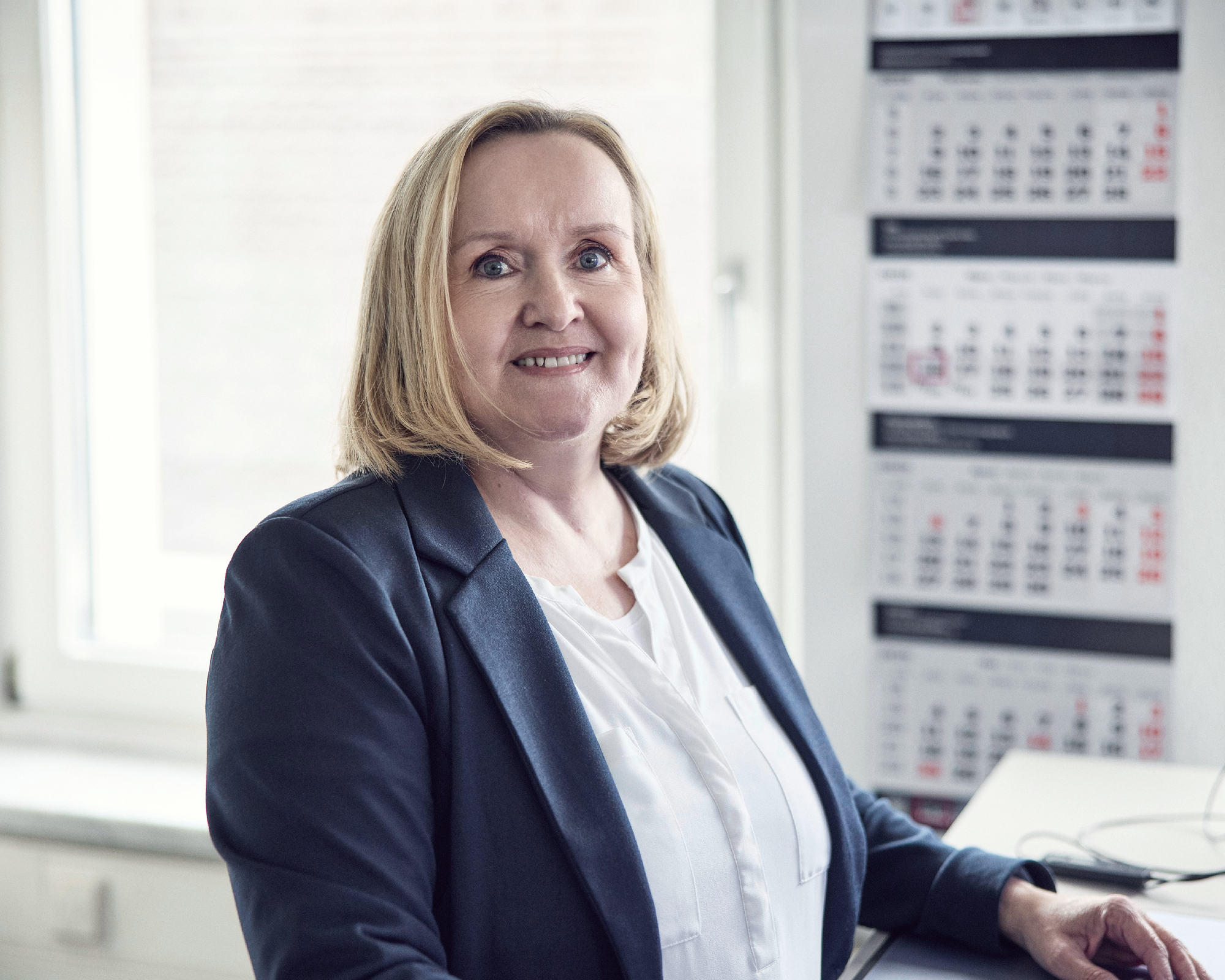 Eine lächelnde Frau mit blonden Haaren steht an einem Schreibtisch in einem hellen Büro, im Hintergrund hängen Wandschalen mit Kalendern.