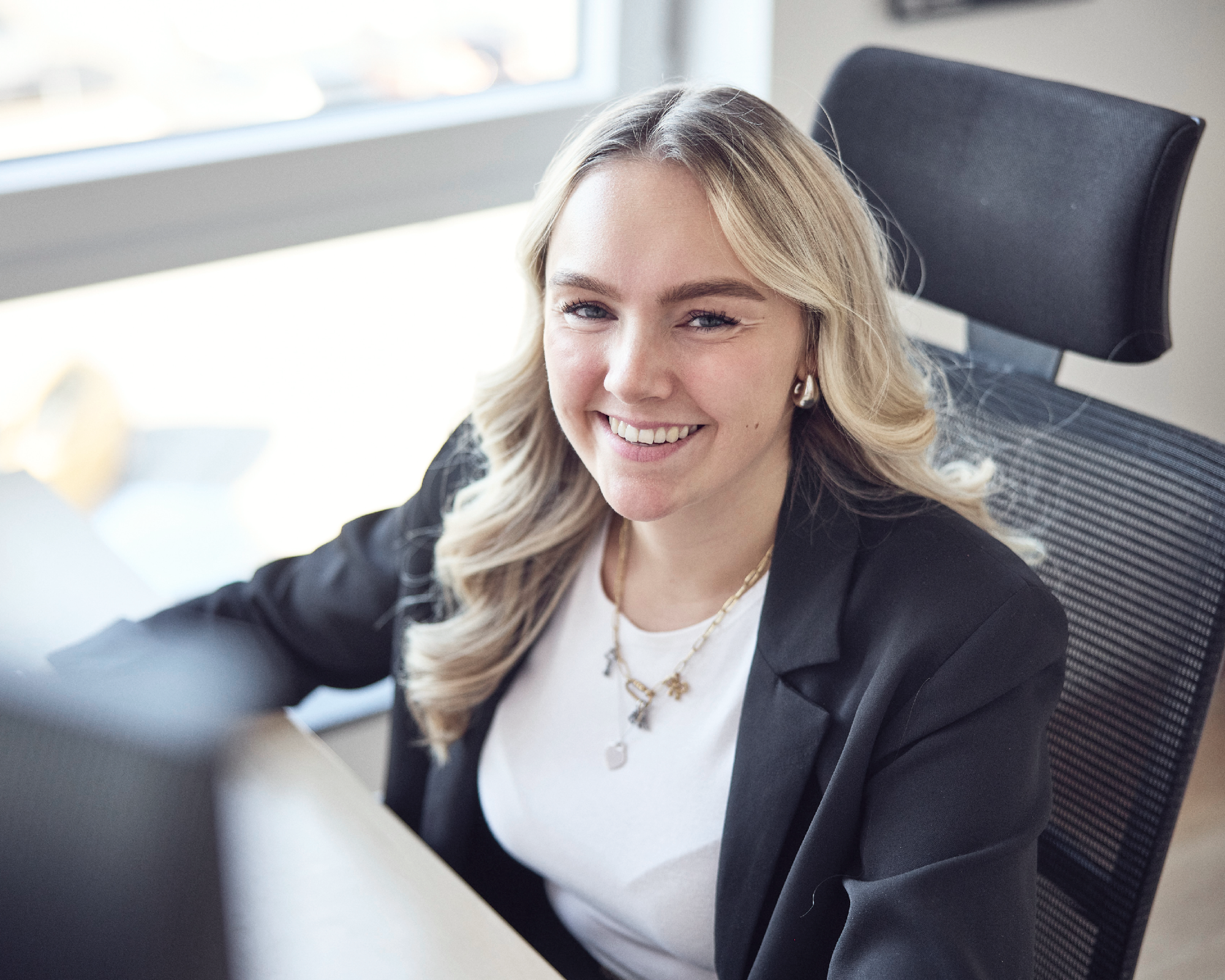 Eine junge Frau mit blonden, lockigen Haaren sitzt an einem Schreibtisch und lächelt freundlich in die Kamera. Sie trägt ein schwarzes Blazer und einen weißen Shirt, während sie vor einem Computerbildschirm sitzt. Im Hintergrund sind Fenster zu sehen, die Tageslicht hereinlassen.