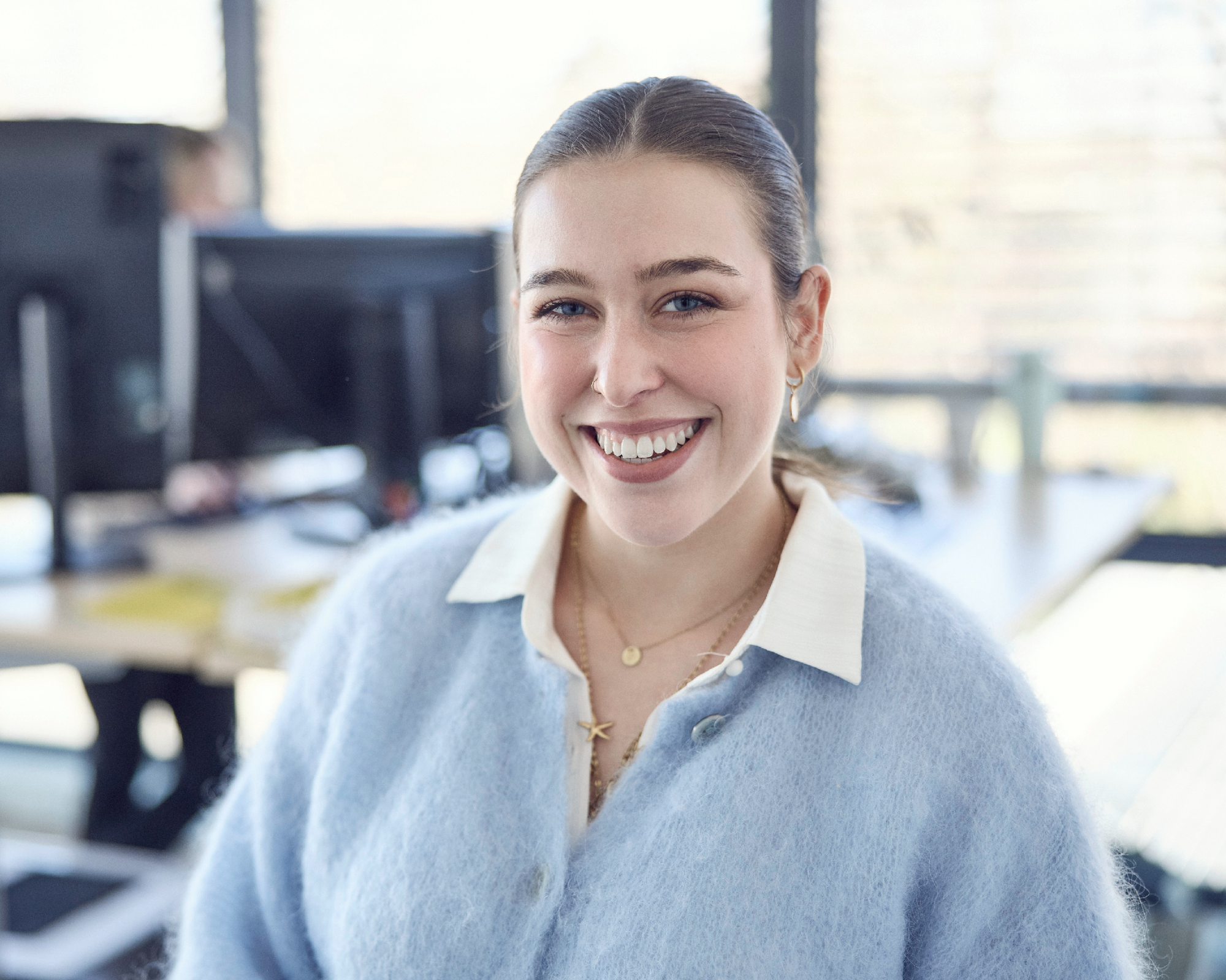 Eine lächelnde junge Frau mit einem hellblauen Pullover und einer weißen Bluse steht in einem modernen Büro vor einem Computerarbeitsplatz.