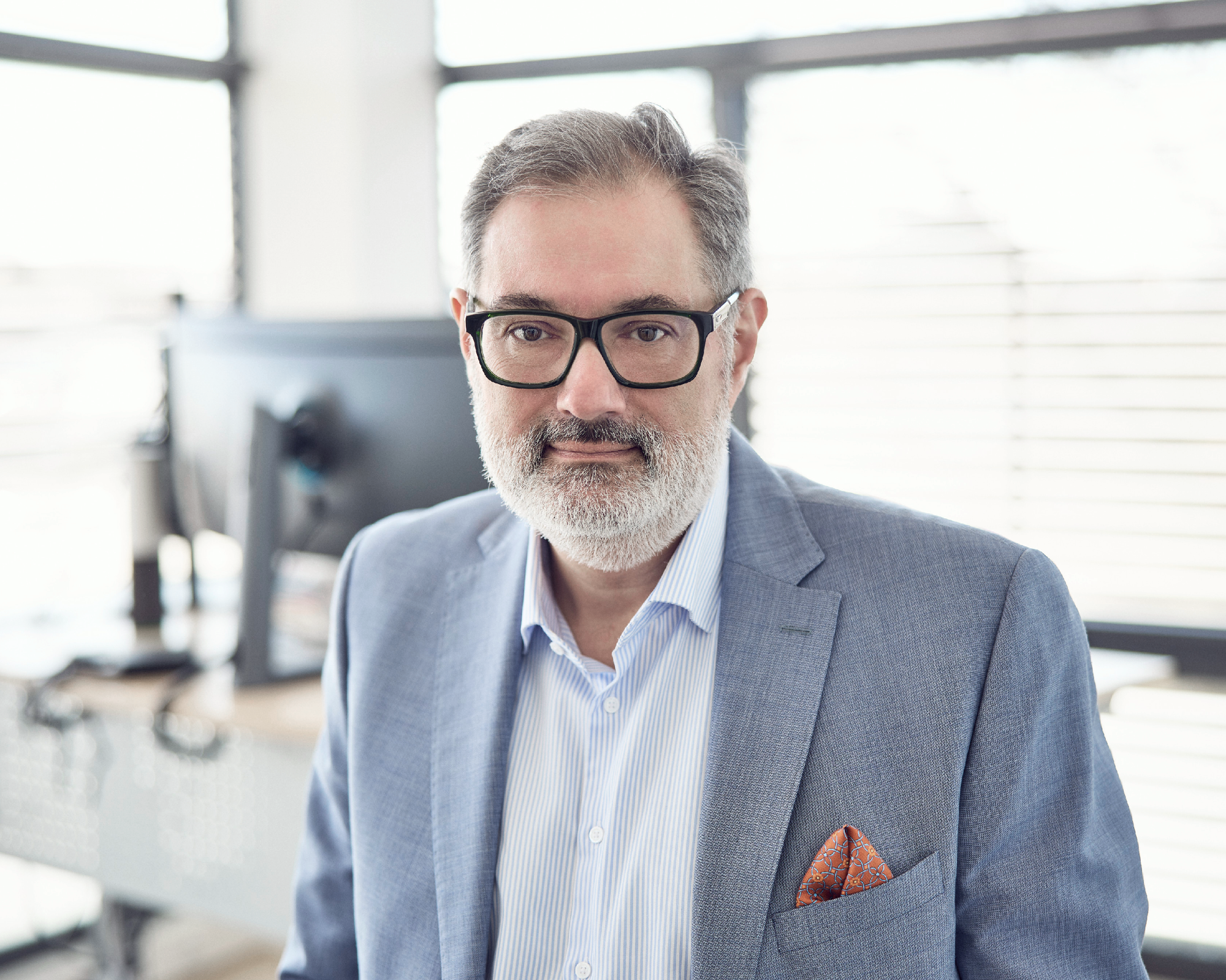 Ein Mann mittleren Alters mit grauem Bart und Brille, trägt einen hellblauen Anzug und ein gestreiftes Hemd, steht in einem modernen Büro mit großen Fenstern im Hintergrund.