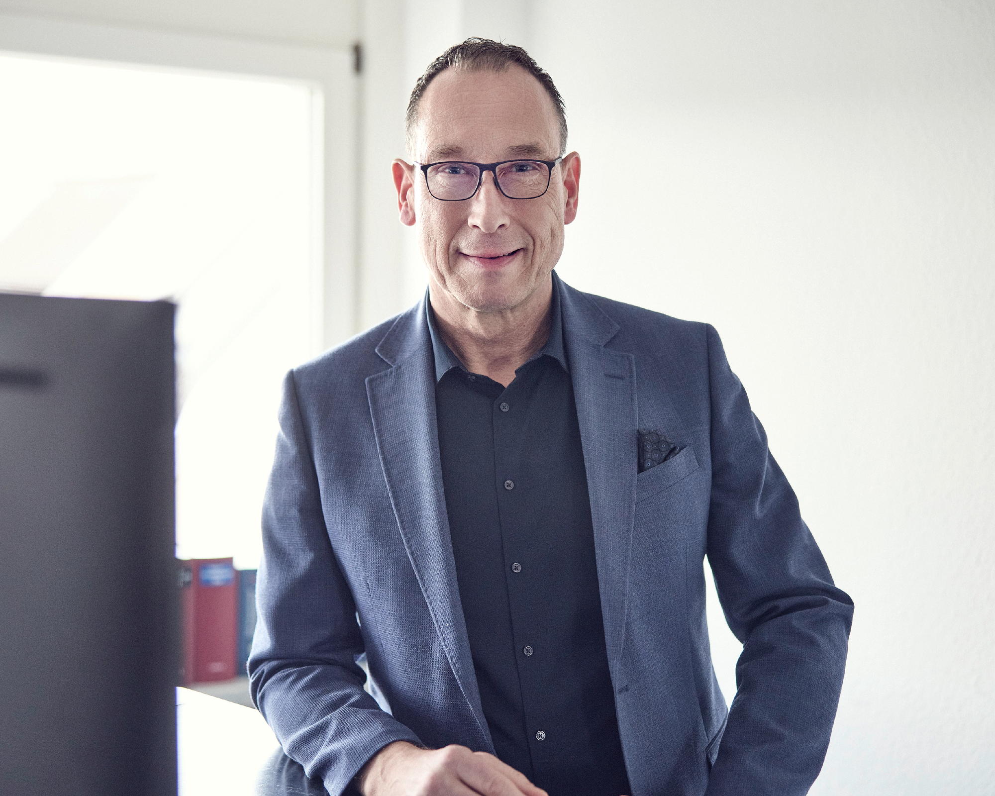 Ein lächelnder Mann in einem eleganten blauen Anzug steht an einem Schreibtisch und schaut in die Kamera, während er von einem Fenster mit natürlichem Licht beleuchtet wird.