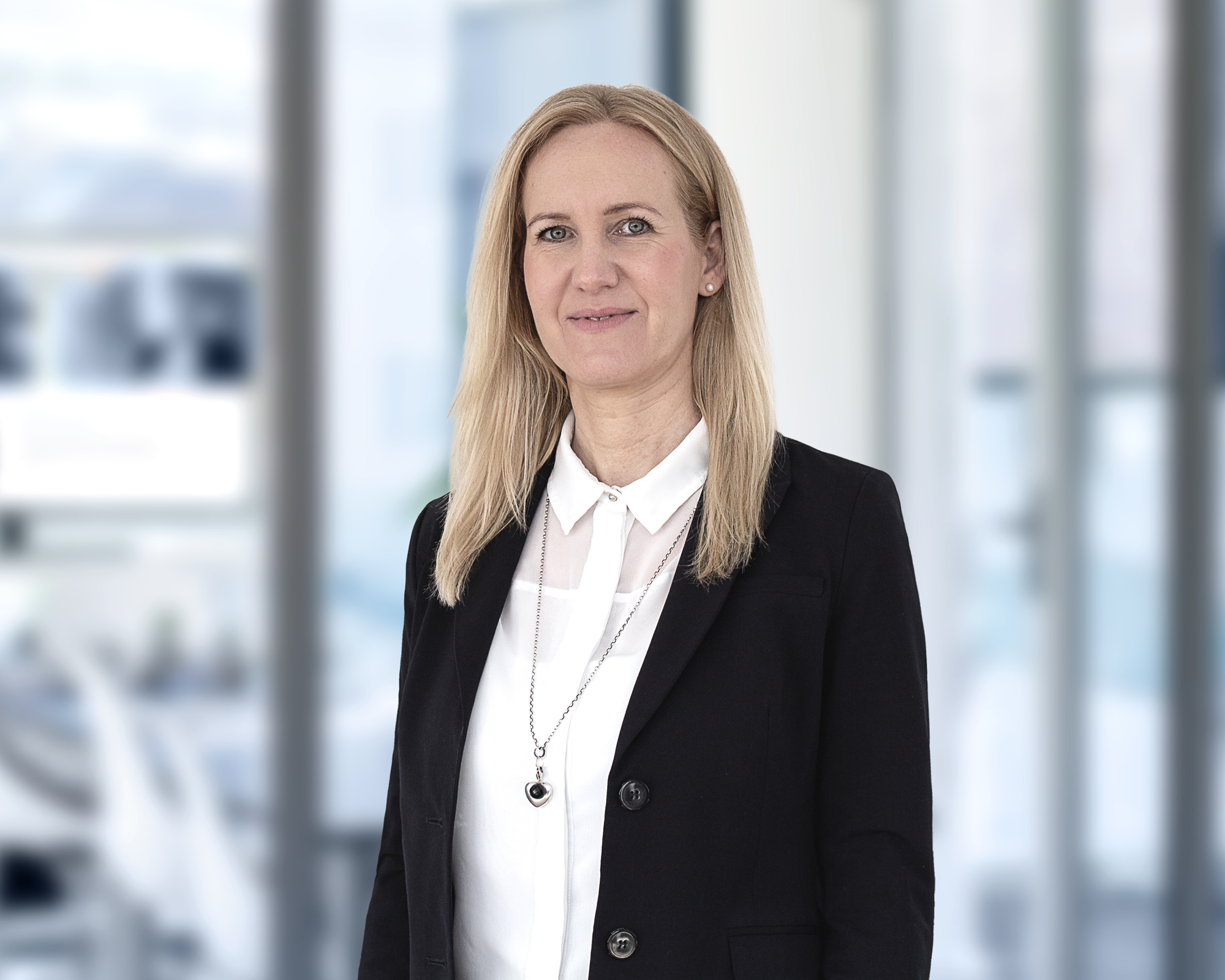 Eine blondhaarige Frau mit mittellanger Haarlänge trägt eine schwarze Jacke über einer weißen Bluse und steht in einem modernen, hellen Büro mit großem Fenster im Hintergrund.