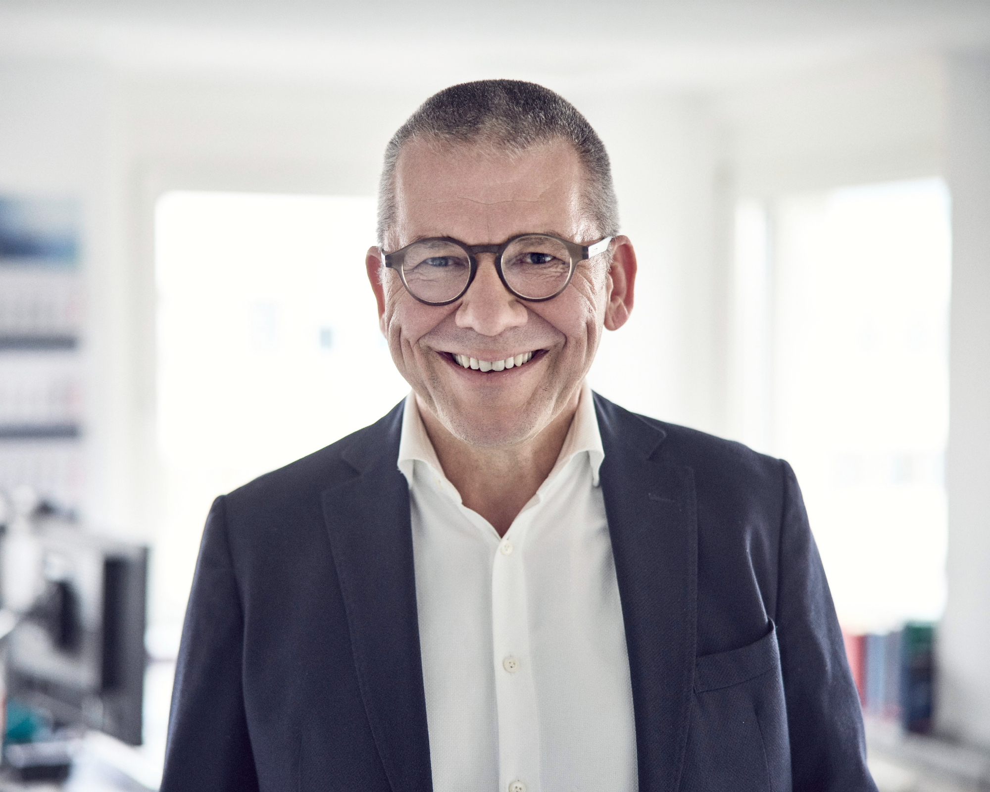 Ein lächelnder Mann mit Brille trägt einen dunklen Anzug und ein weißes Hemd, steht in einem hellen Raum mit Büromöbeln im Hintergrund.