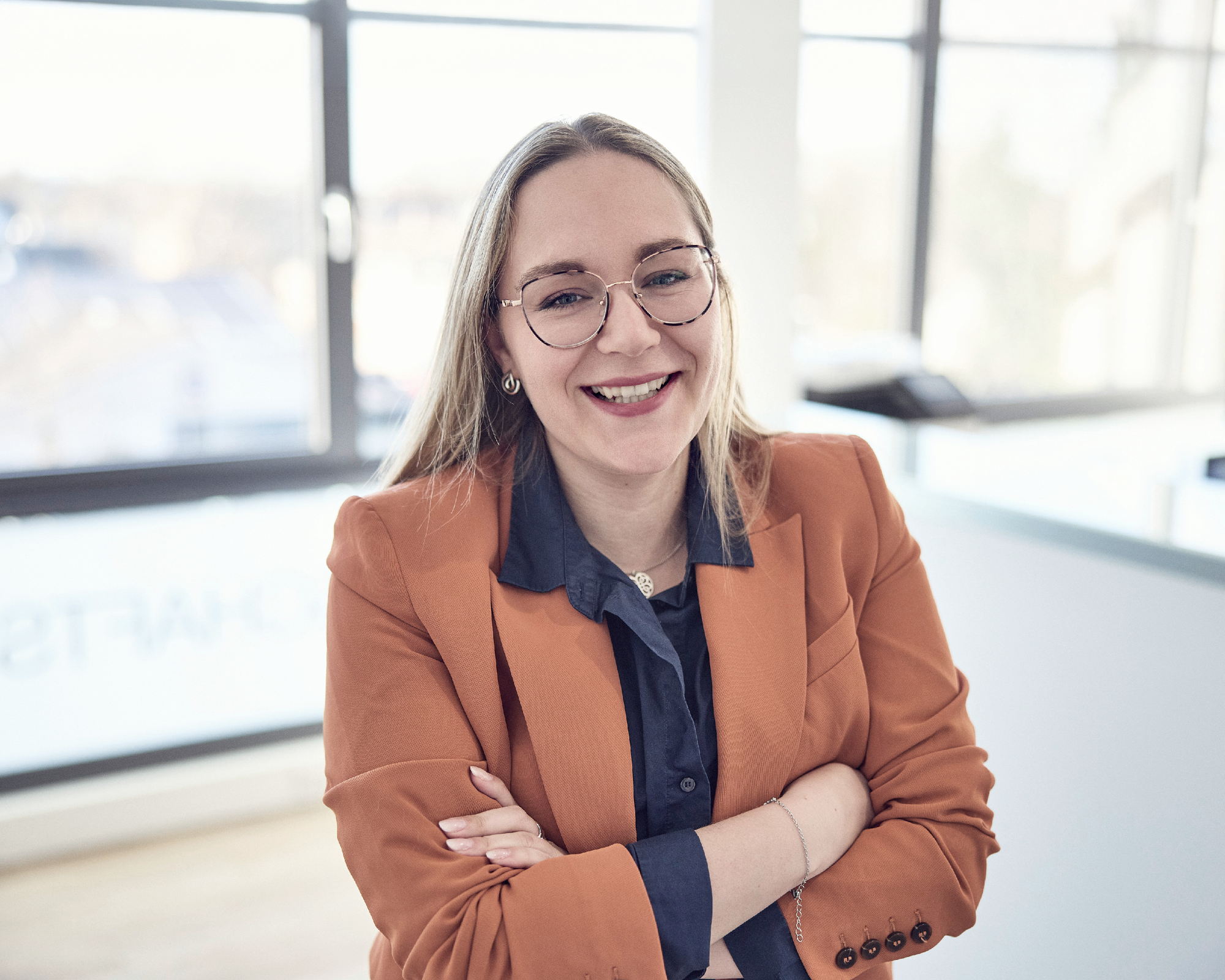 Eine Frau steht mit verschränkten Armen vor großen Fenstern und lächelt. Sie trägt ein orangefarbenes Blazer-Jacket und eine blaue Bluse. Im Hintergrund sind verschwommene Büroelemente zu erkennen.