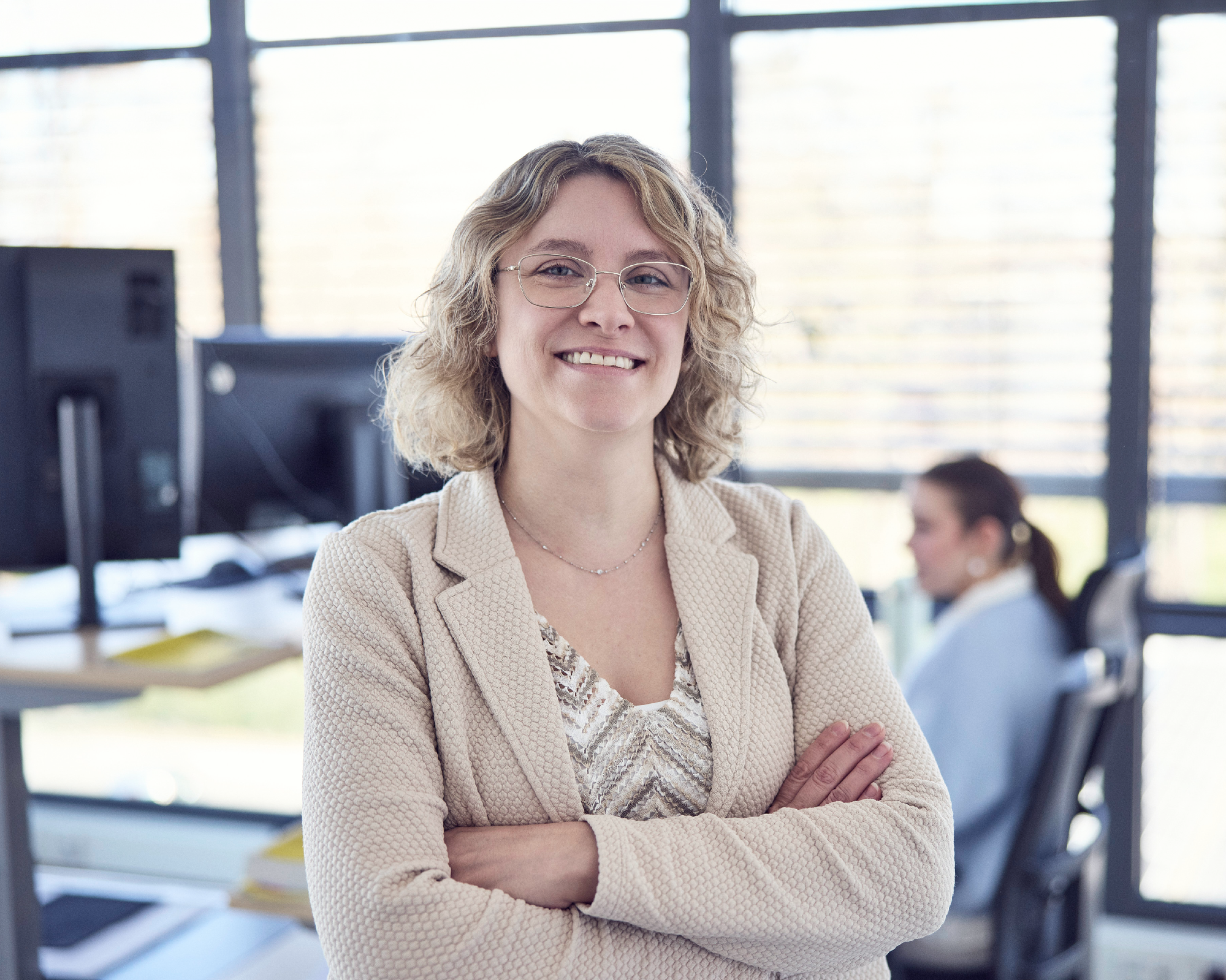 Eine lächelnde Frau mit lockigem, blondem Haar und Brille steht selbstbewusst mit verschränkten Armen in einem modernen Büro, im Hintergrund sind zwei weitere Mitarbeiterinnen zu sehen.
