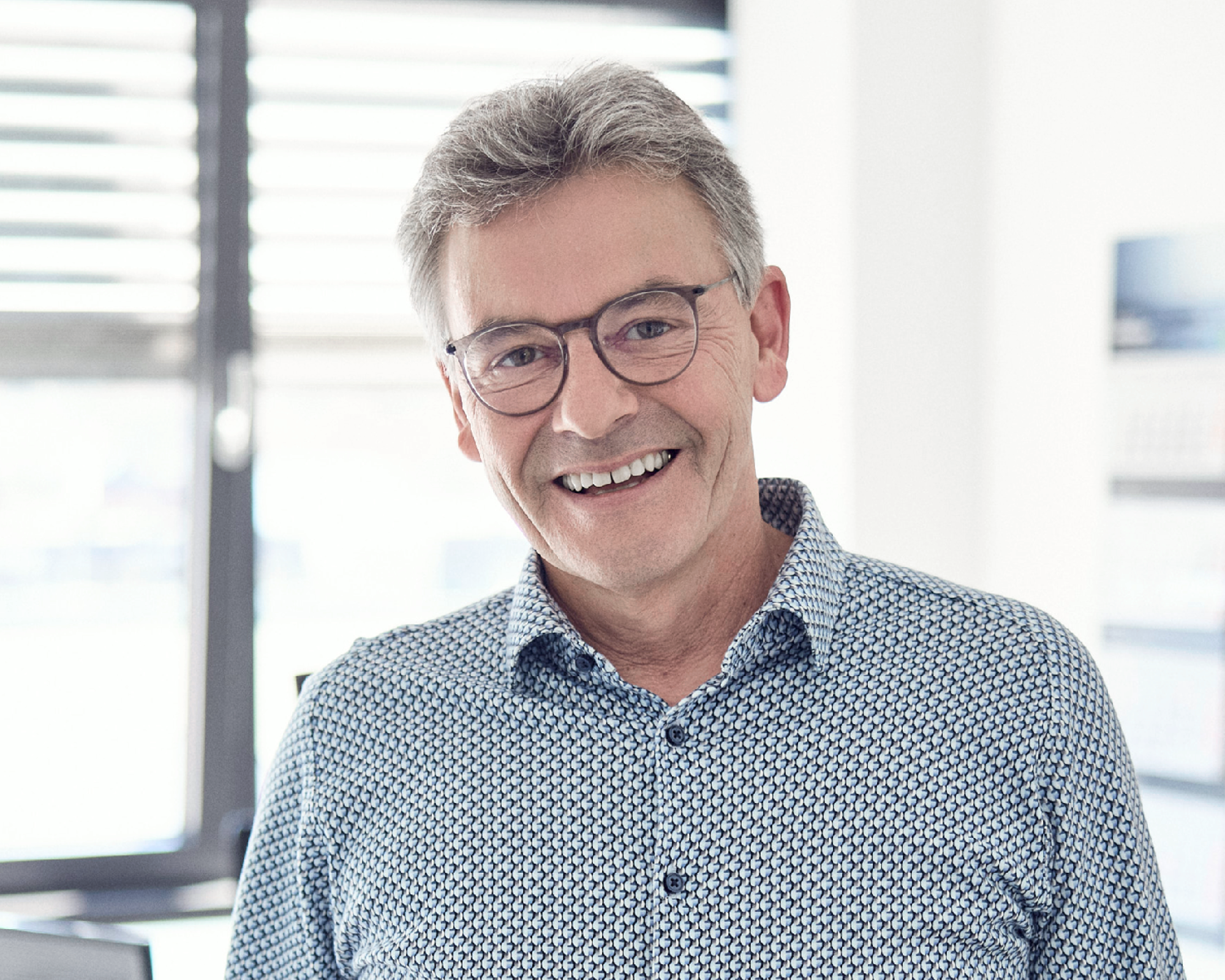 Ein lächelnder Mann mit grauen Haaren und Brille trägt ein hellblaues Hemd mit Muster und steht in einem hellen, modernen Büro.