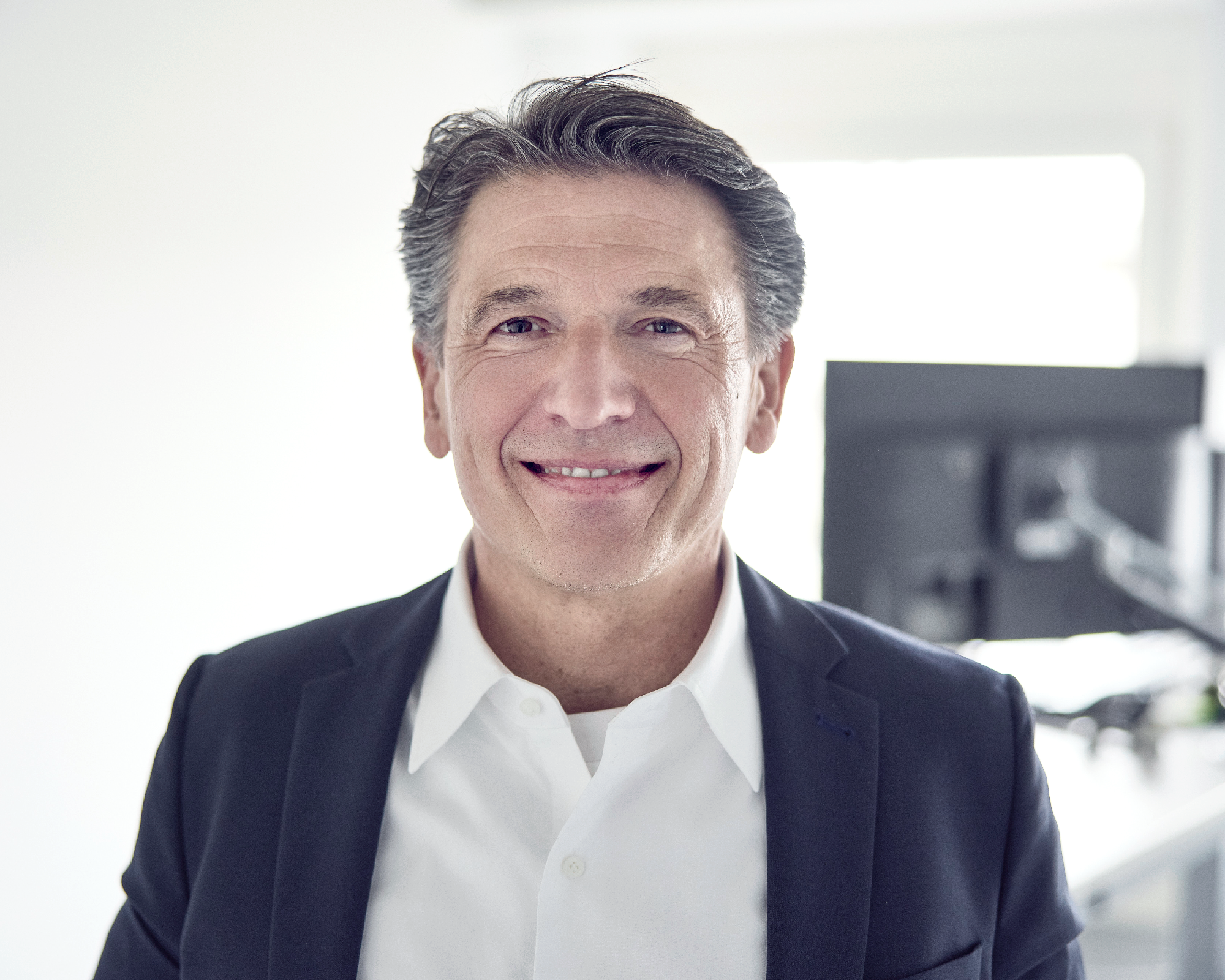 Ein Mann mittleren Alters mit kurzen, grauen Haaren lächelt freundlich in einem modernen Büro, das einen Schreibtisch und einen Computer im Hintergrund zeigt.