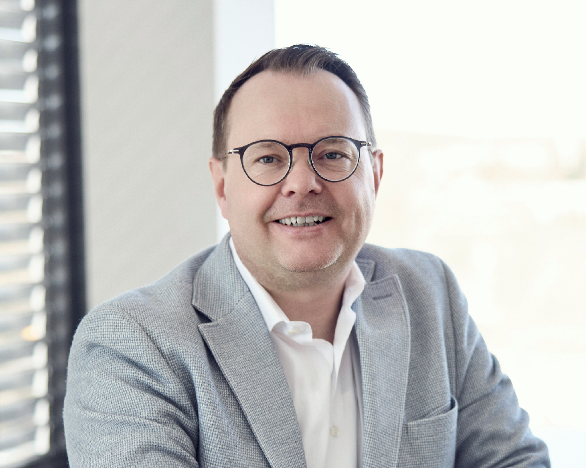 Ein lächelnder Mann mit Brille trägt einen hellgrauen Blazer und ein weißes Hemd, während er in einem gut beleuchteten Raum sitzt.