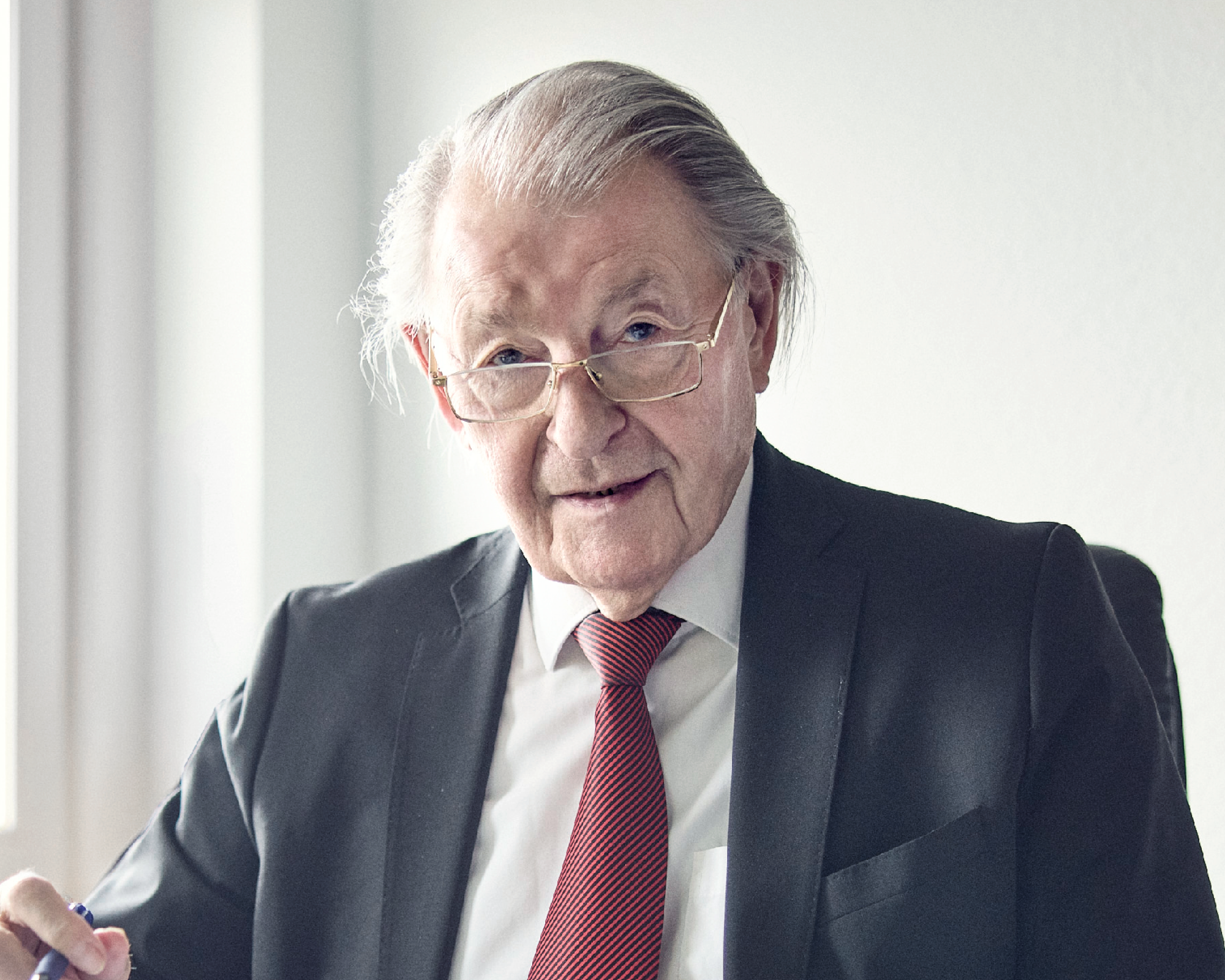 Ein älterer Mann mit schütterem grauem Haar und einer Brille, trägt einen schwarzen Anzug und eine rot gemusterte Krawatte. Er sitzt an einem Tisch und schaut direkt in die Kamera, während er einen Stift hält. Die Umgebung wirkt modern und hell.