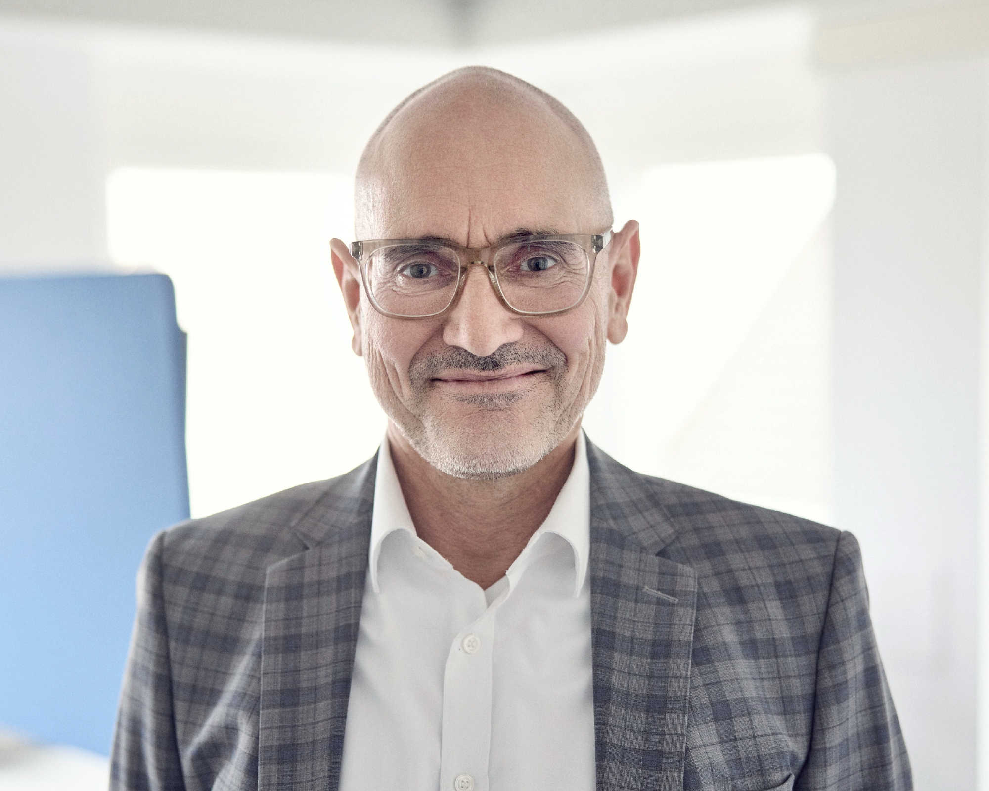 Ein lächelnder, älterer Mann mit Brille trägt einen karierten Anzug und ein weißes Hemd und steht in einem hellen Büro.