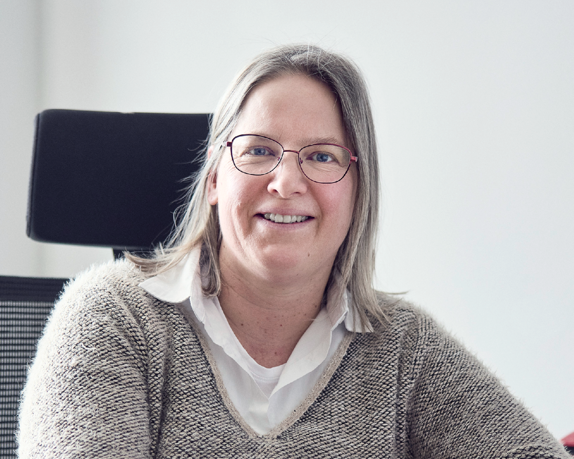 Eine lächelnde Frau mit langen, grauen Haaren und Brille sitzt an einem Schreibtisch, trägt einen hellen Pullover und schaut freundlich in die Kamera.