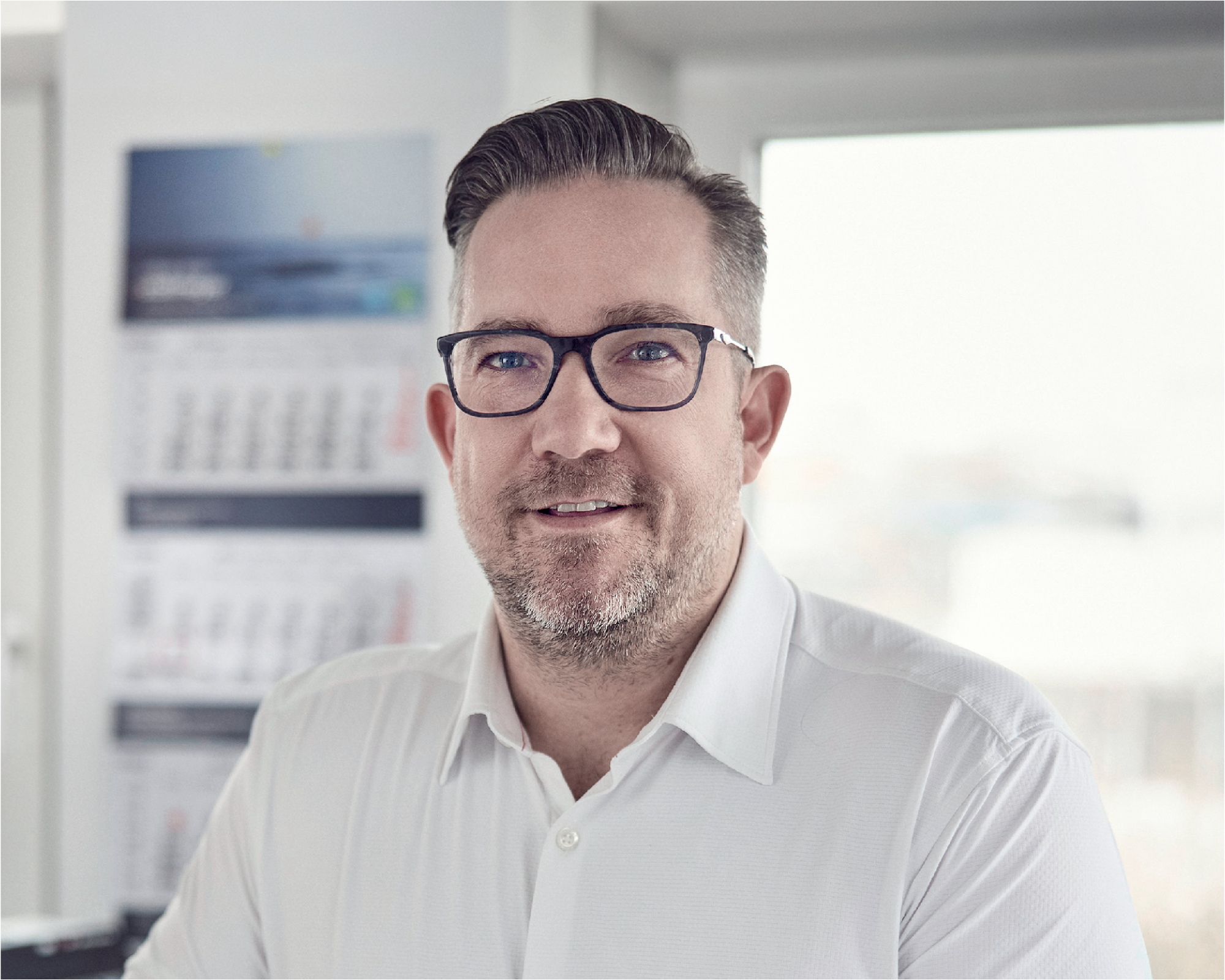 Ein lächelnder Mann mit Brille trägt ein weißes Hemd und steht in einem modernen Büro mit Fenstern, im Hintergrund sind Kalender und Dokumente sichtbar.