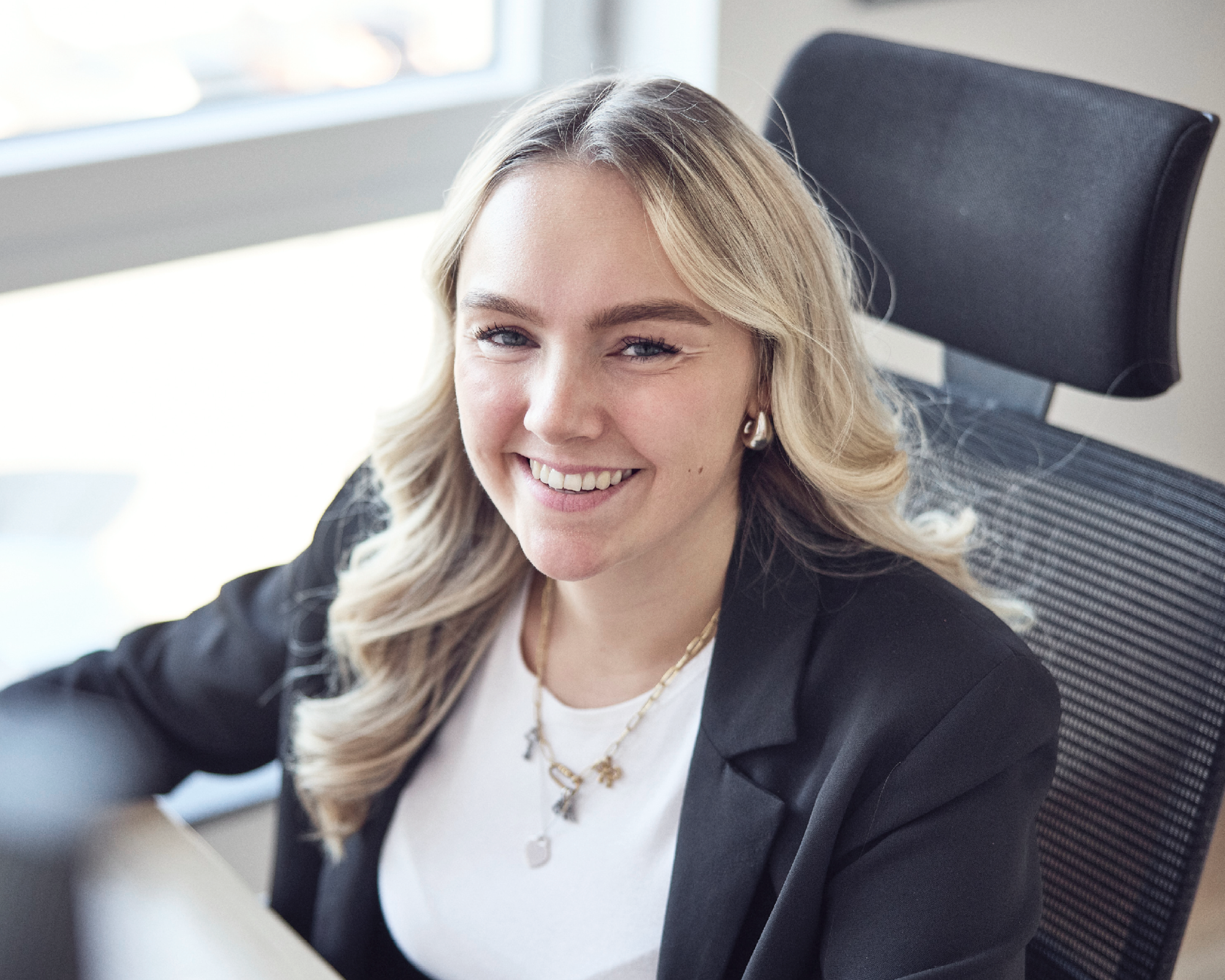 Eine lächelnde Frau mit langen, blonden Haaren sitzt an einem Schreibtisch in einem modernen Büro und trägt ein schwarzes Blazer.
