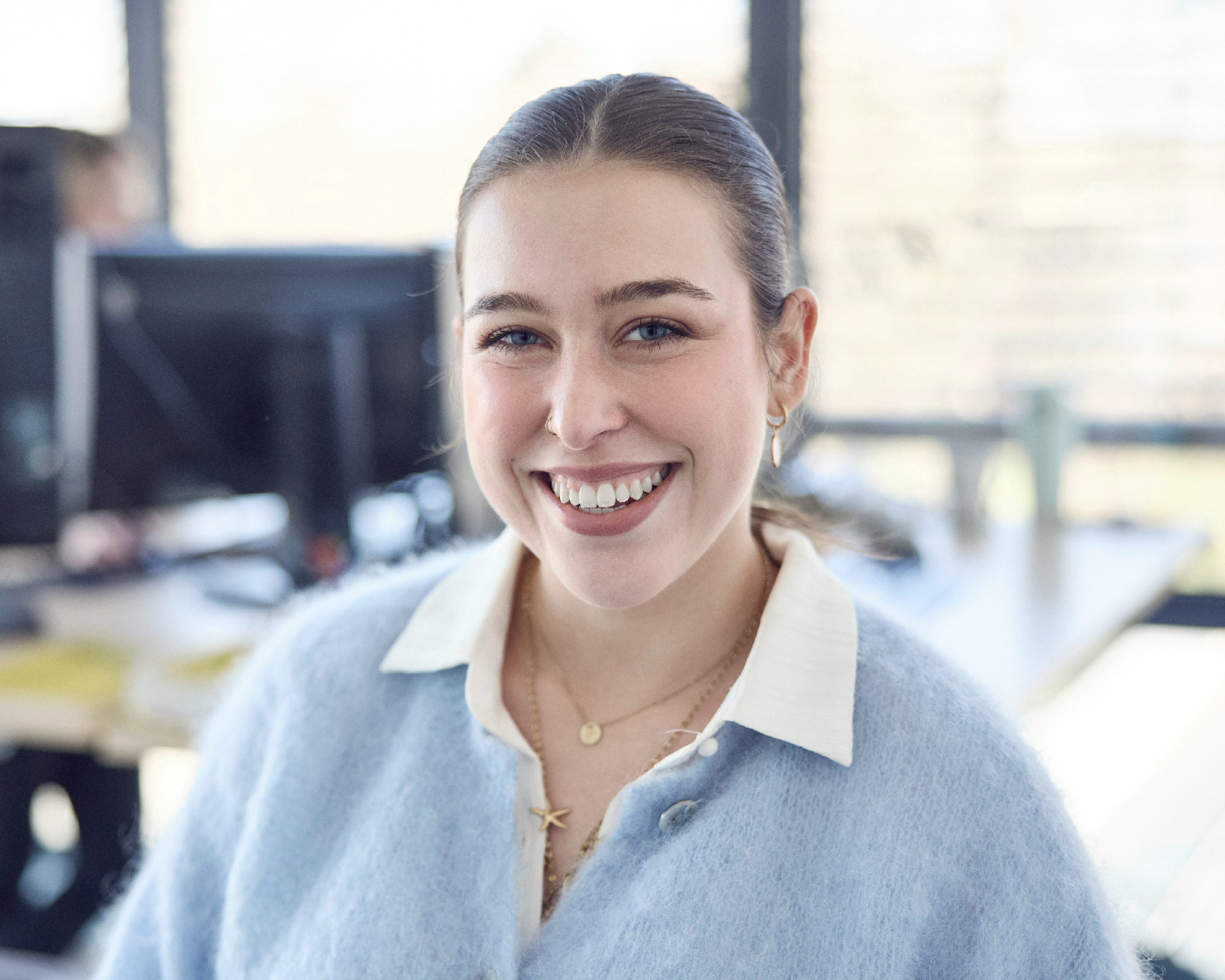 Eine lächelnde junge Frau mit braunen Haaren und blauer Strickjacke sitzt in einem modernen Büro, während sie in die Kamera schaut. Im Hintergrund sind mehrere Computerarbeitsplätze sichtbar.