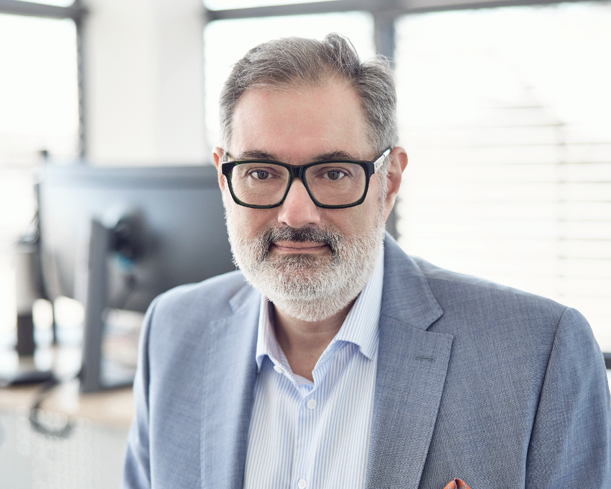 Ein Mann mit Brille und grauem Bart trägt einen hellen Anzug und ein gestreiftes Hemd. Er schaut direkt in die Kamera, während im Hintergrund ein moderner Arbeitsplatz mit Computern sichtbar ist.
