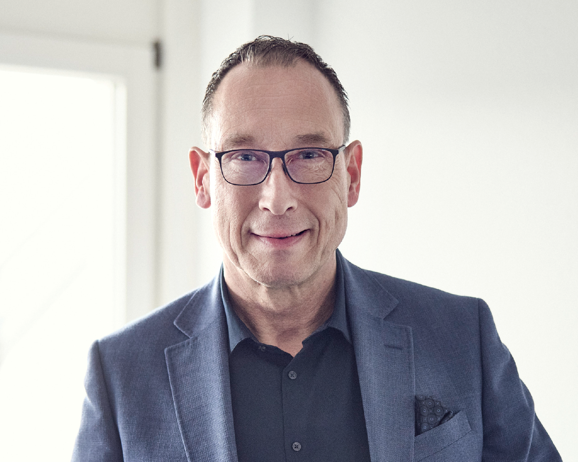 Ein Mann mit Brille und kürzeren, dunklen Haaren trägt einen blauen Blazer und steht vor einem Fenster, während er freundlich in die Kamera lächelt.