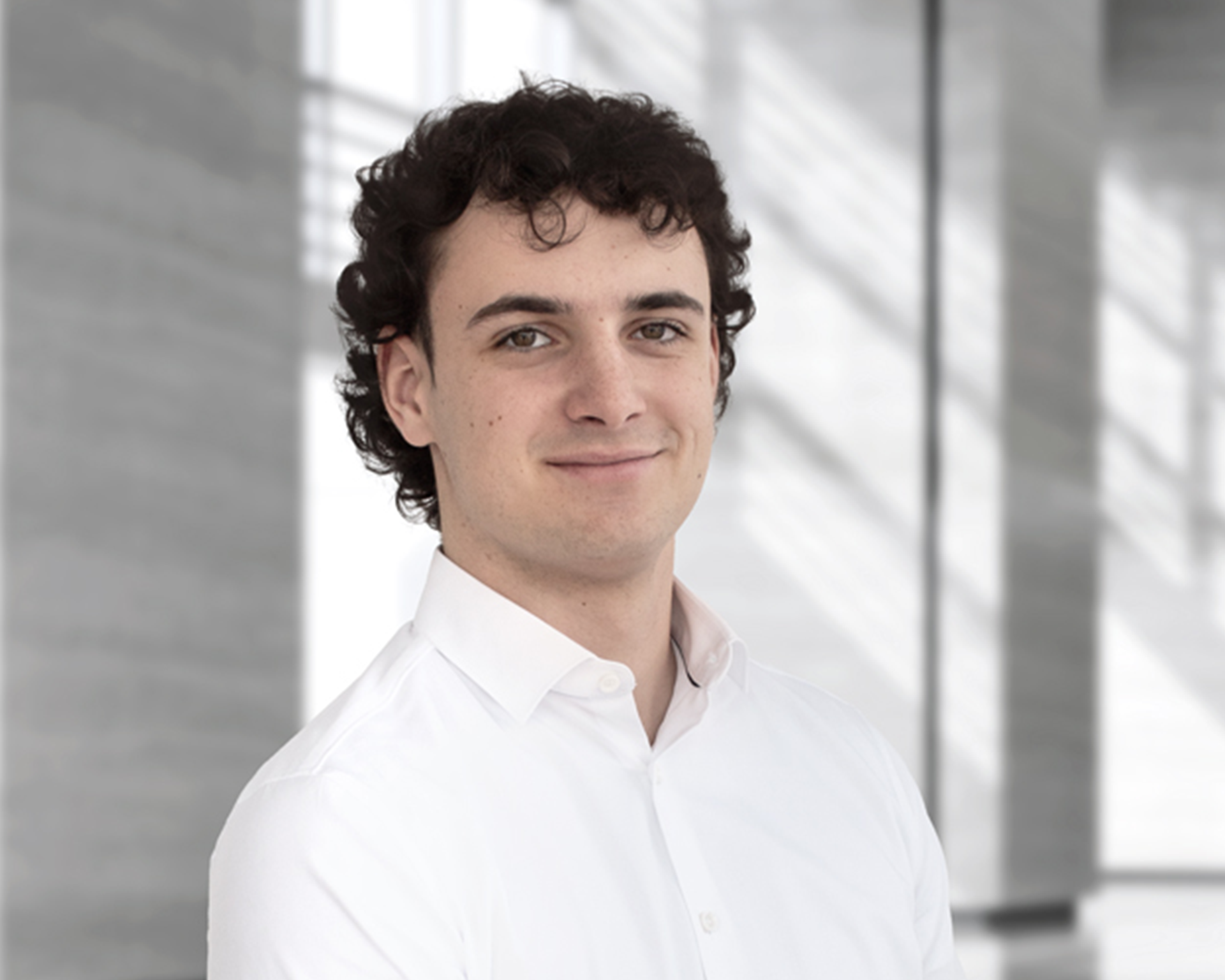 Ein junger Mann mit lockigem Haar trägt ein weißes Hemd und steht in einem modernen, hellen Innenraum mit großen Fenstern.
