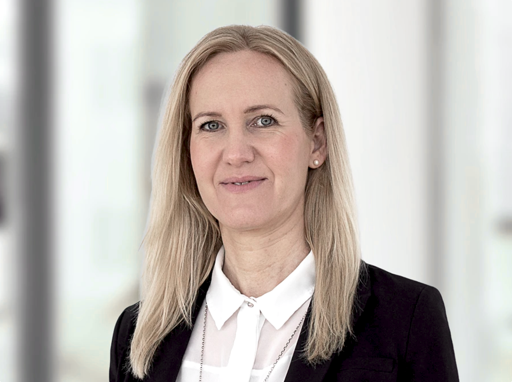Eine blonde Frau mit langen Haaren trägt ein schwarzes Blazer und ein weißes Hemd, lächelt in die Kamera und steht vor einem unscharfen, hellen Hintergrund.