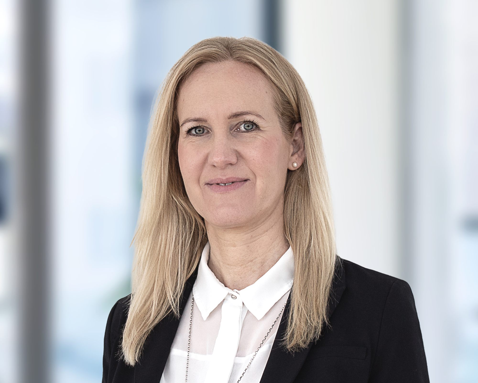 Eine blonde Frau mit langen Haaren trägt eine schwarze Blazerjacke und ein weißes Hemd, lächelt freundlich in die Kamera. Der Hintergrund ist unscharf und hell.