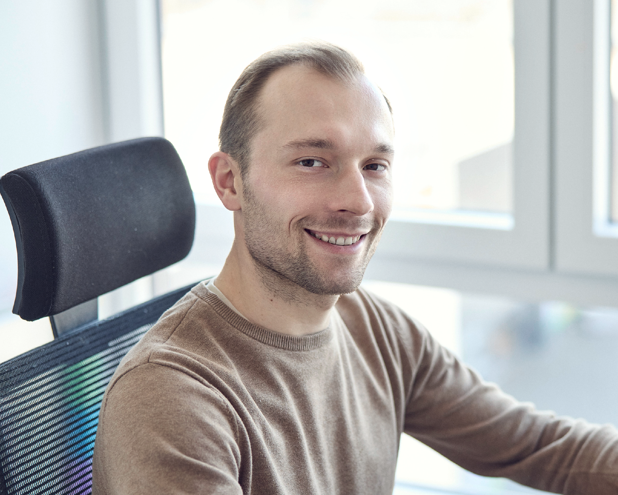Ein Mann in einem hellbraunen Pullover lächelt freundlich in die Kamera, während er in einem modernen Bürostuhl sitzt, mit großen Fenstern und hellem Licht im Hintergrund.
