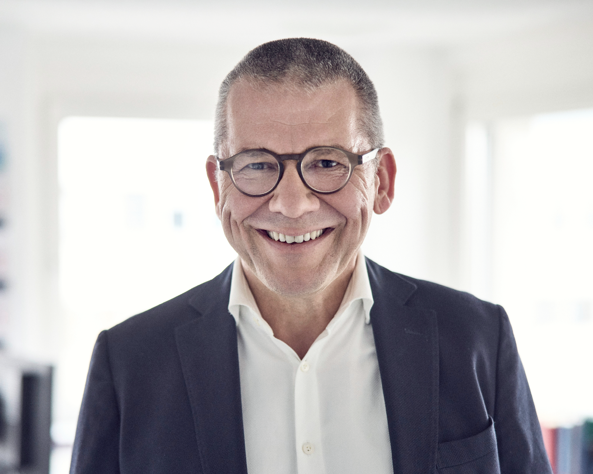 Ein lächelnder Mann mit kurzen, grauen Haaren und Brille trägt einen dunklen Blazer und ein weißes Hemd. Er steht in einem hellen Raum mit unscharfen Möbeln im Hintergrund.