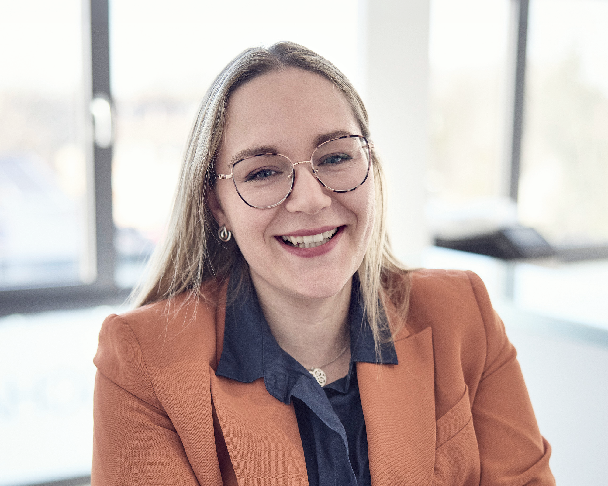 Eine lächelnde Frau mit langen, glatten Haaren sitzt in einem modernen Büro. Sie trägt eine orangefarbene Blazerjacke und eine blaue Bluse, während sie durch ihre Brille blickt. Im Hintergrund sind hell erleuchtete Fenster zu sehen.