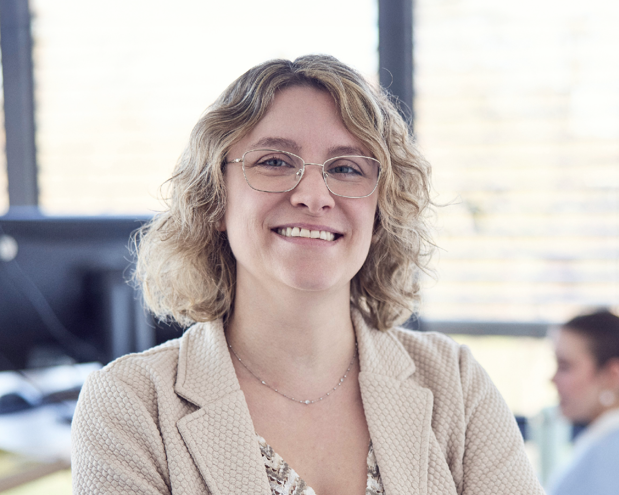 Eine lächelnde Frau mit lockigem, blondem Haar und Brille steht im Vordergrund eines modernen Büros, während im Hintergrund weitere Mitarbeitende zu sehen sind.