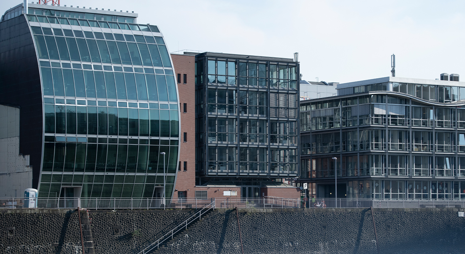 Moderne Gebäude mit unterschiedlichen architektonischen Stilen, darunter große Glasfronten und gewellte Fassaden, stehen entlang eines Ufers.