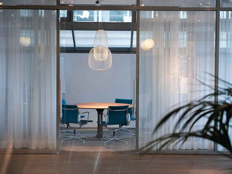 Ein moderner Konferenzraum mit einem runden Holztisch und stühlen in Aqua-Tönen, erhellt von einer stylischen Pendelleuchte. Der Raum ist durch transparente Vorhänge vom Rest des Büros abgetrennt und hat eine ruhige Atmosphäre.