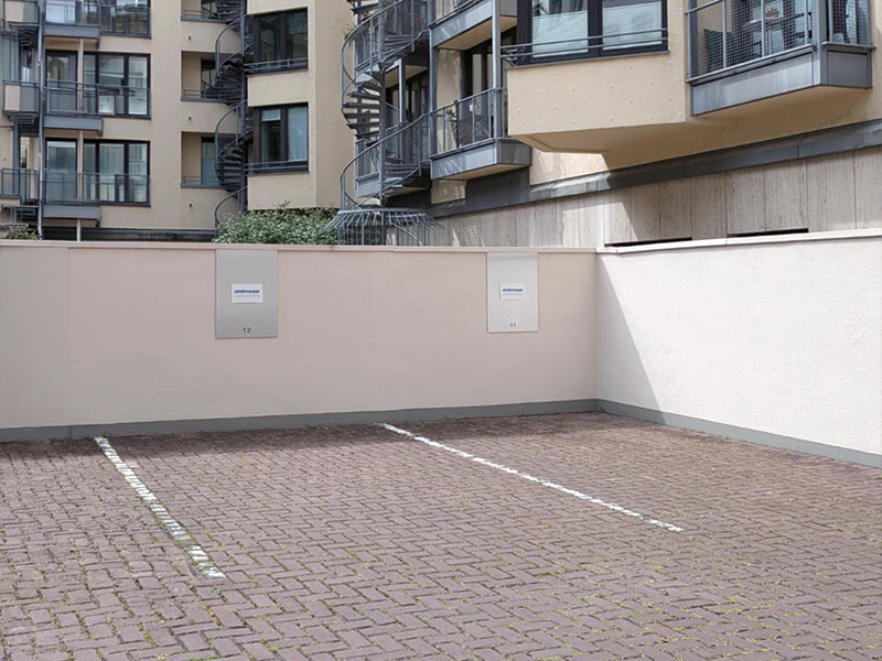 Leerer Parkplatz mit grauer Pflasterung, umgeben von hellen Gebäuden mit Balkonen und einer Wendeltreppe im Hintergrund. An der Wand sind zwei graue Kästen angebracht.